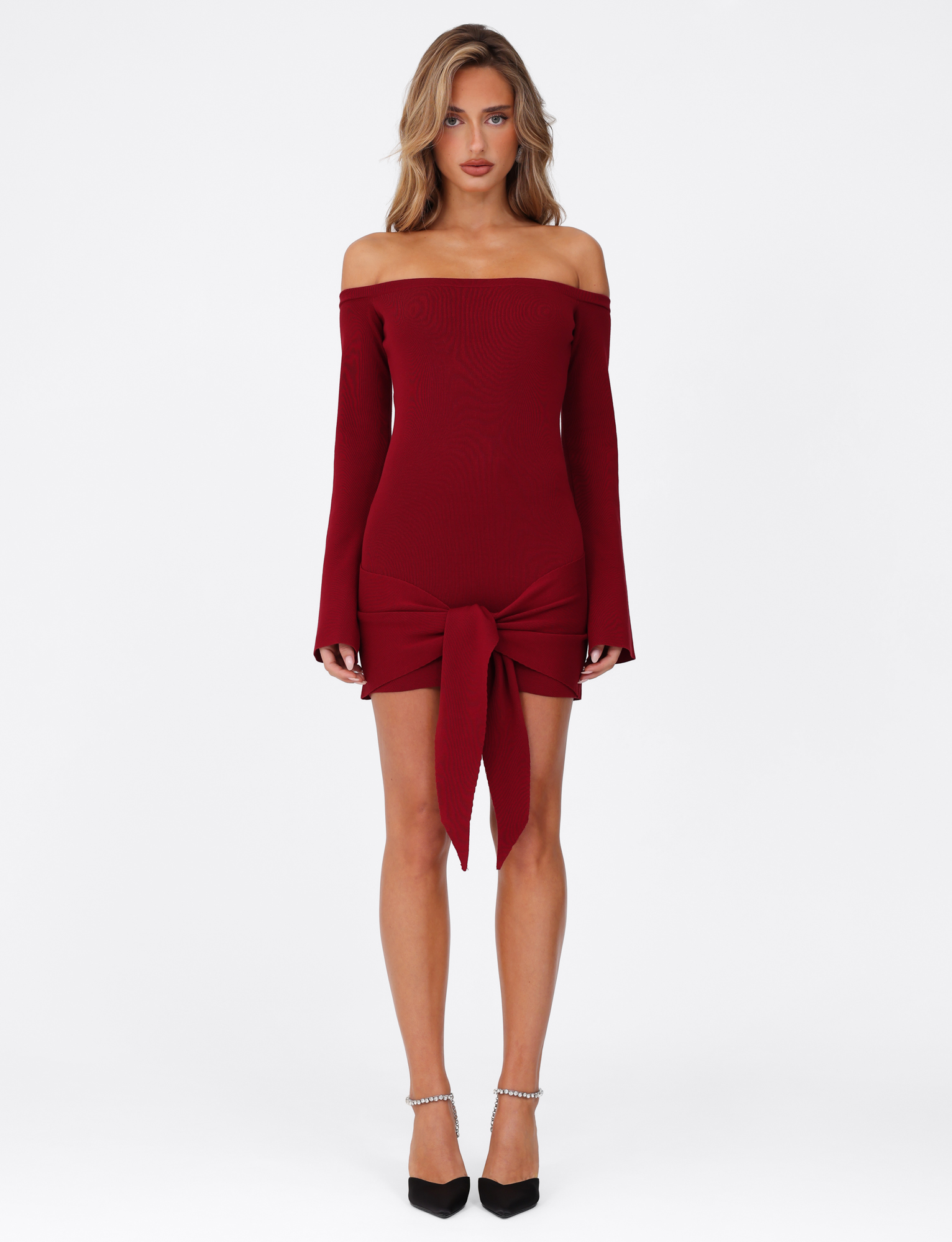 A woman faces forward in the TIGERMIST CYPRESS MINI DRESS - RED : WINE, a bodycon off-shoulder mini with long sleeves and a large front-tie. She pairs it with black ankle-strap heels and loose wavy hair, posed against a white background.