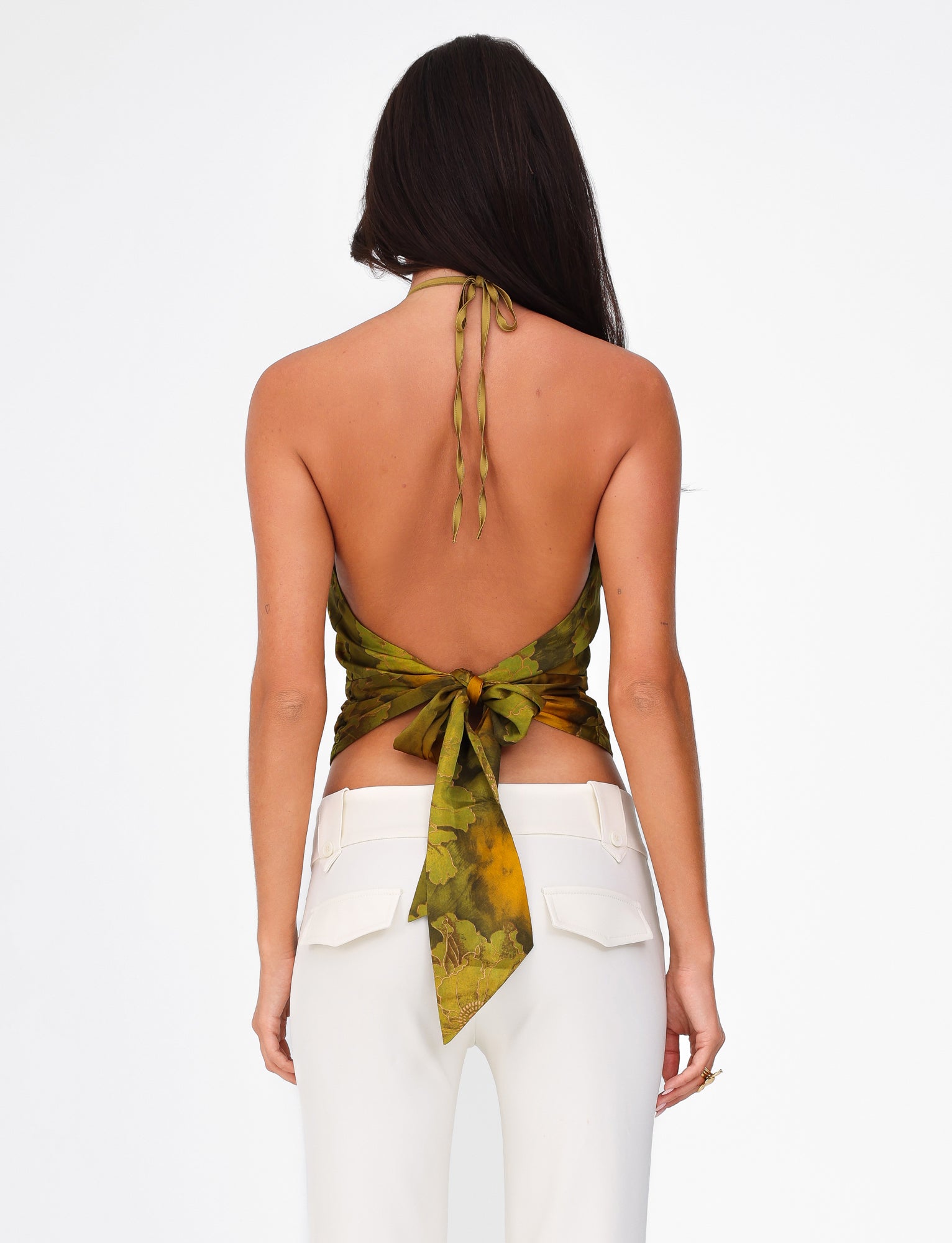 Rewritten Sentence: Rear view of a person with long dark hair wearing the TIGERMIST MONROE TOP - GREEN : FLORAL and white high-waisted pants. The halter neckline top is tied at the back, revealing their bare back, while the pants feature flap pockets with buttons. The background is plain white.