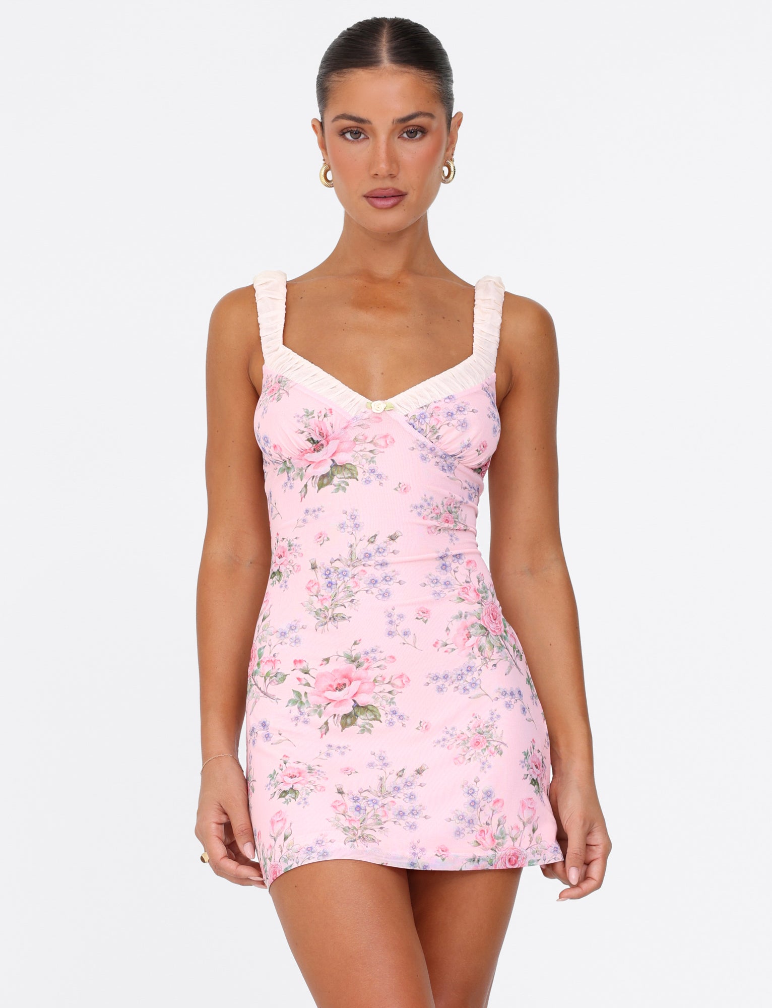 Someone wearing the Amaran Mini Dress by Tigermist, a pink mini dress with a wild flower floral print, poses against a plain background. The dress showcases a V-neckline and ruffled straps. The individual has long hair tied back and is accessorized with earrings.