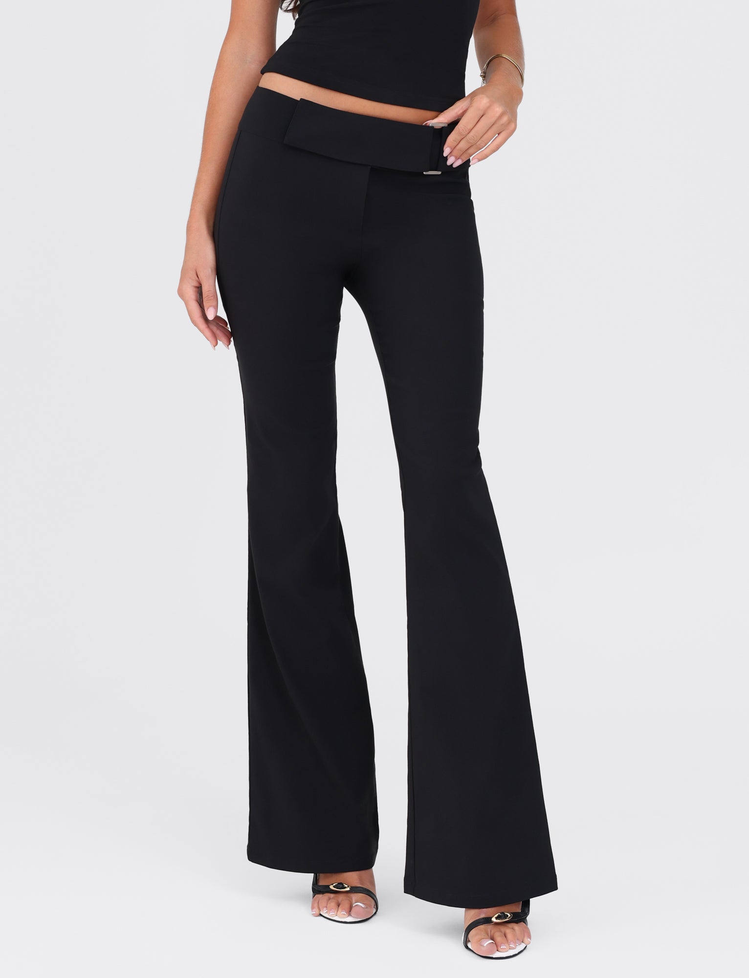 A person is wearing the VESPER PANT - BLACK by TIGERMIST, showcasing black flared pants with a fitted silhouette and a cropped black top. Their left hand rests by their side, while their right hand lightly touches their hip. The image captures only the lower half of the person, from the waist down to their feet, which are clad in open-toed shoes.
