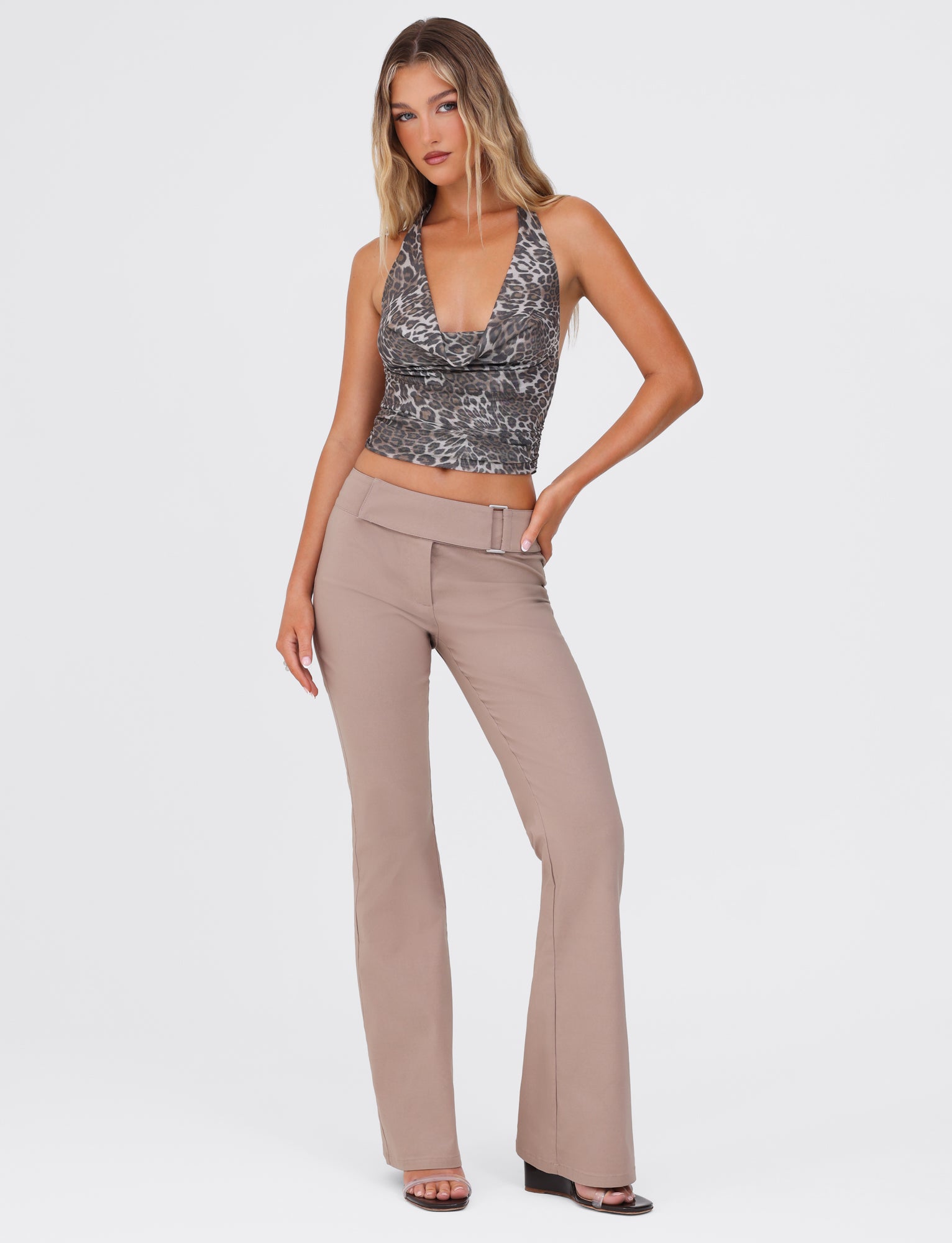 A woman with long blonde hair is posing against a white background. She is wearing a patterned sleeveless top with a deep V-neckline and the VESPER PANT - BROWN : TAN by TIGERMIST, featuring a fitted silhouette and low-rise waist. Her left hand rests on her hip, and she is standing in black open-toe heels.