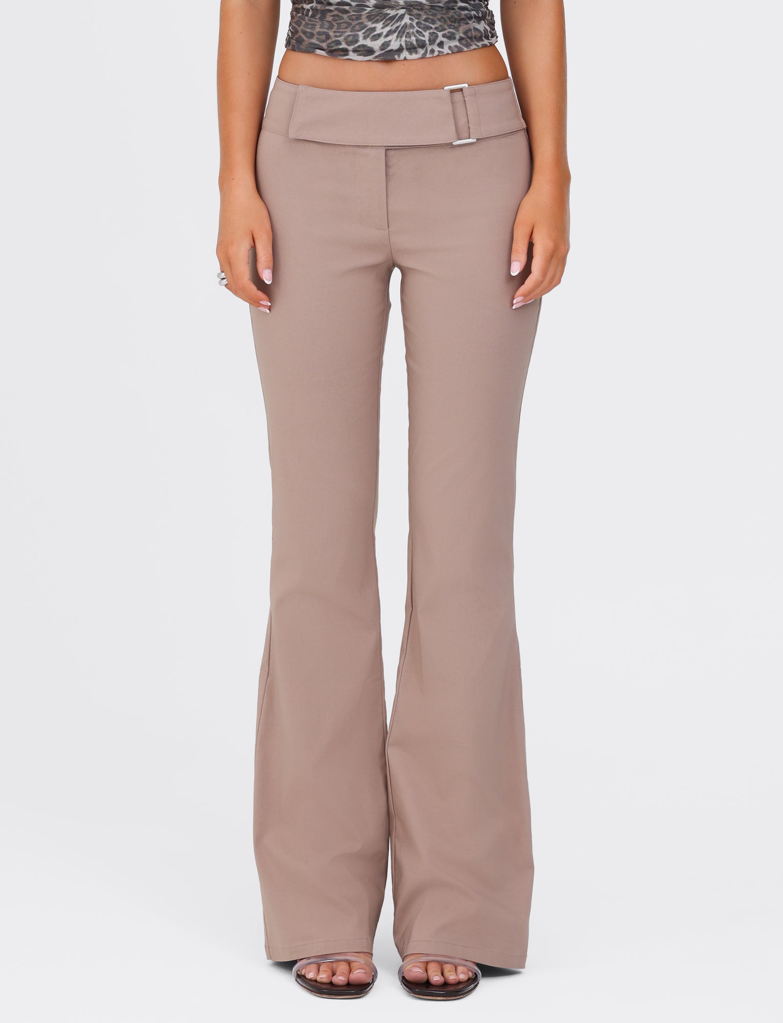A person wearing the VESPER PANT in Brown Tan by TIGERMIST, featuring flared legs, a high waist, and a belt buckle detail. Paired with an animal print top, the individual stands with their hands relaxed at their sides, exuding a polished yet casual style.