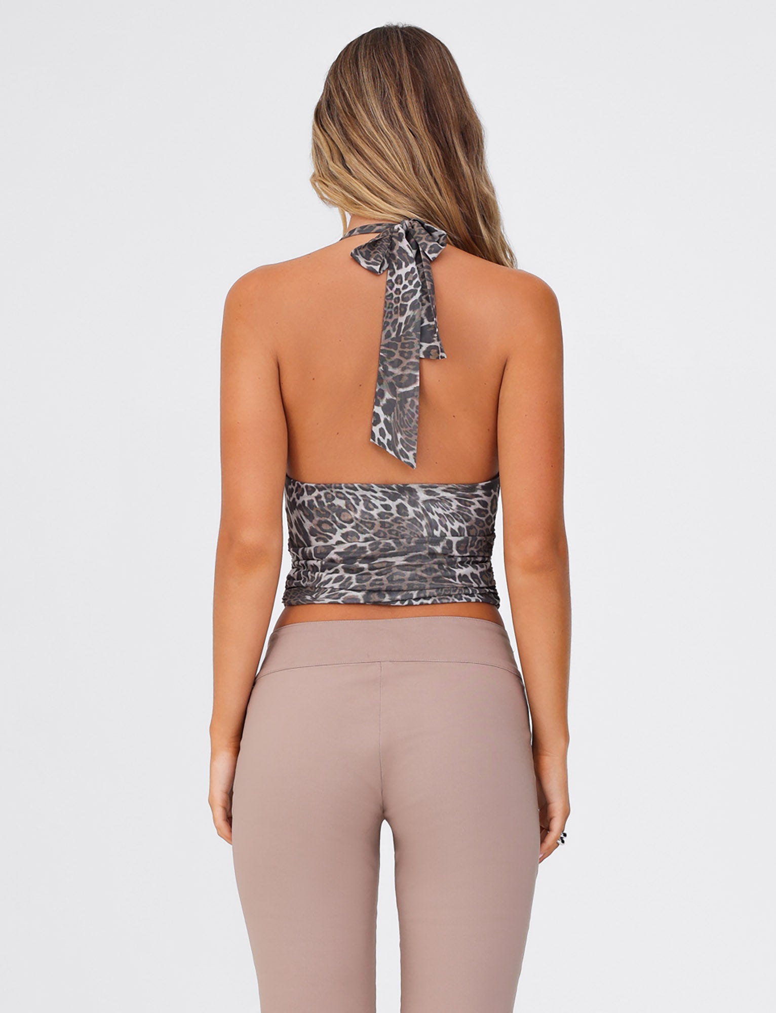 A woman with long, wavy hair stands with her back to the camera. She is wearing the TIGERMIST RILYNN TOP - NEUTRAL : LEOPARD, a halter top that ties behind her neck and features a leopard print design. She pairs it with high-waisted, fitted taupe pants made from high stretch fabric, all set against a plain white background.