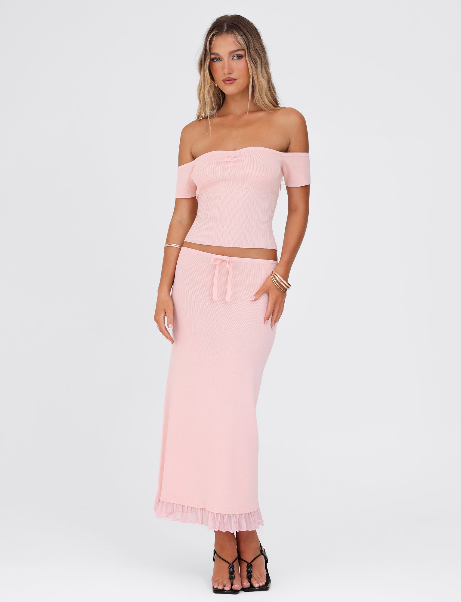 A woman models the TIGERMIST LILYANNA TOP in baby pink, paired with a matching ankle-length skirt, against a plain white backdrop. She completes her look with bracelets on her right wrist and black strappy heels, and her hair cascades in loose waves.