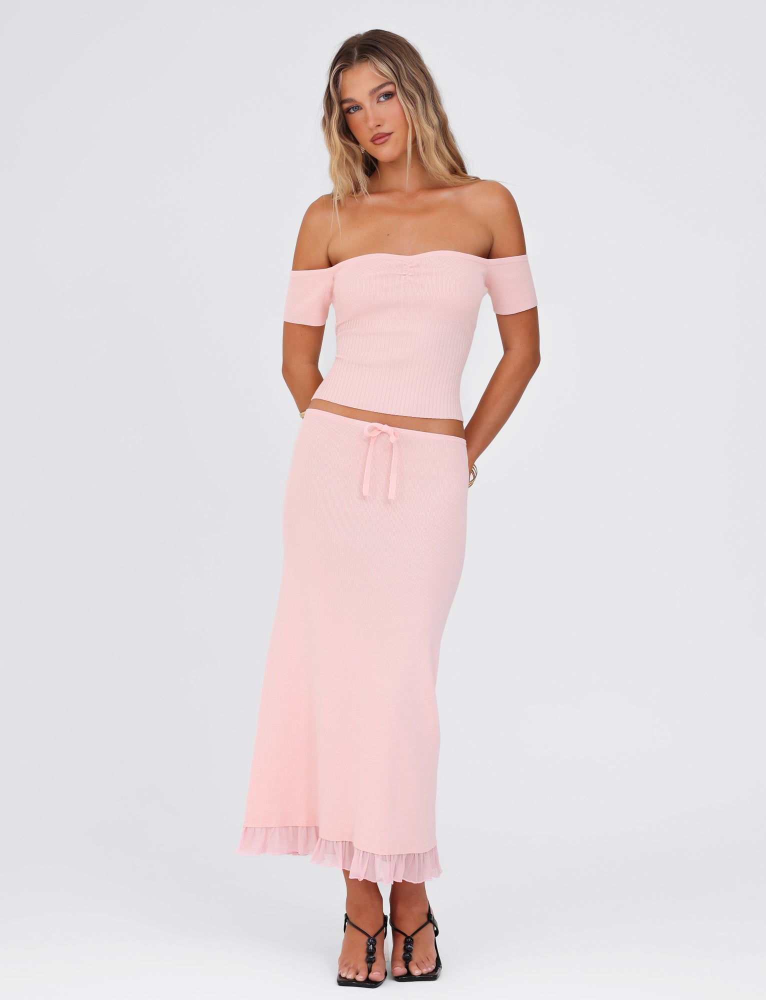 A woman stands against a plain background, modeling the TIGERMIST LILYANNA SKIRT in a soft baby pink shade. She complements the relaxed fit with a light pink off-shoulder top and black strappy heels. Her long, wavy hair cascades down her shoulders as she confidently places one hand on her hip.