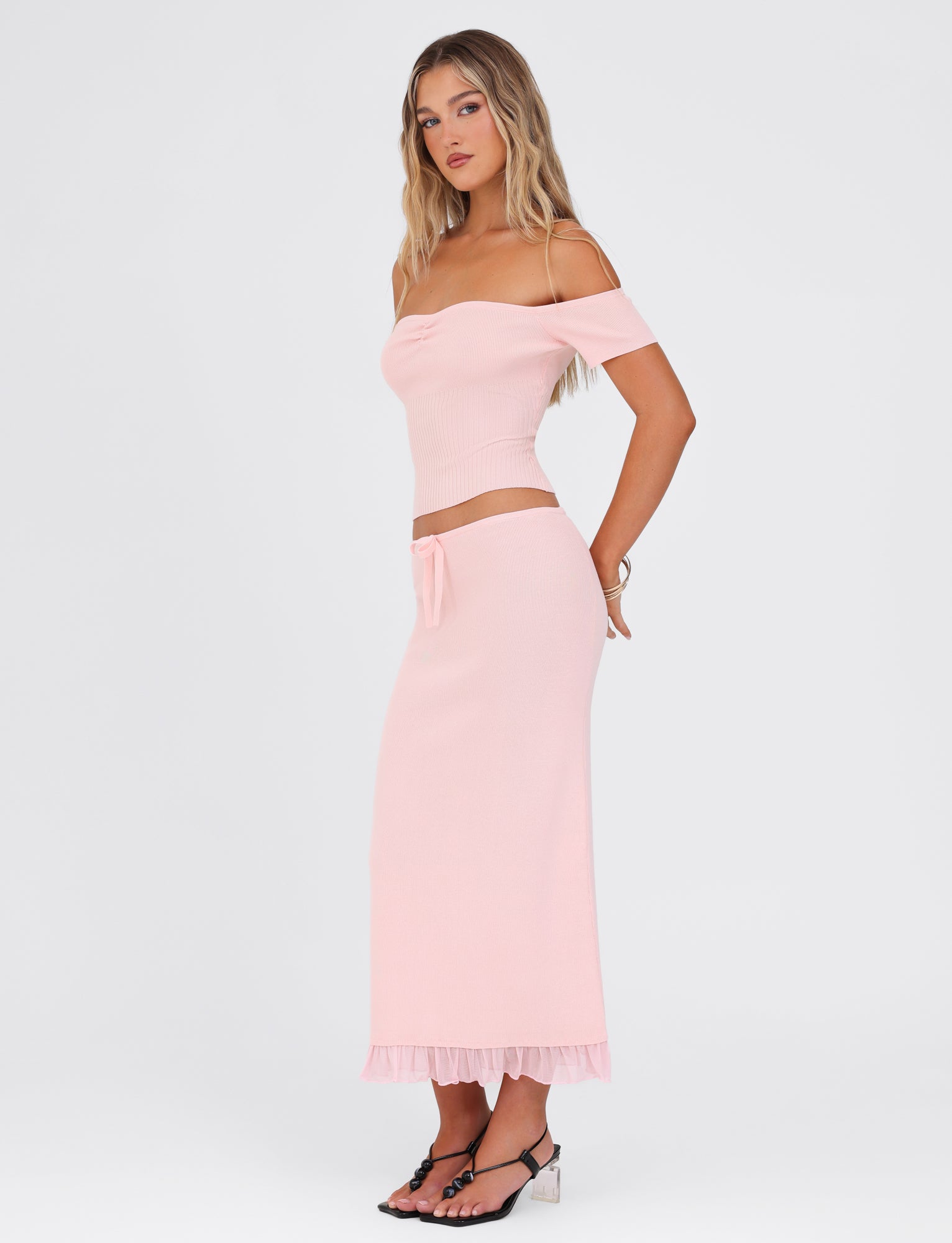 A person wearing the TIGERMIST LILYANNA SKIRT in baby pink with a matching off-the-shoulder top stands against a white background. They are wearing black sandals and have long, wavy hair. The chic ensemble is showcased as they stand slightly turned to the side.