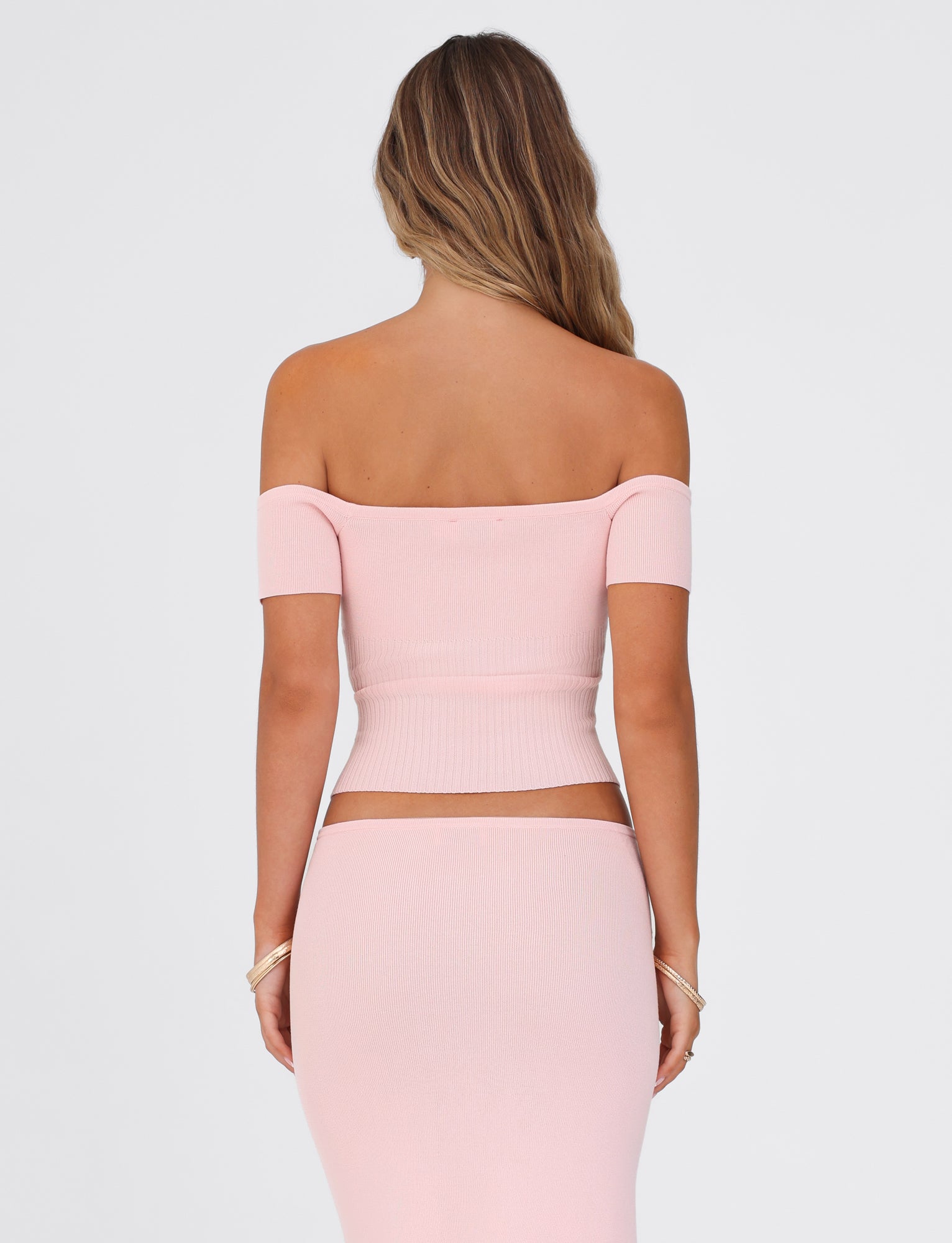 A woman with long hair is wearing the TIGERMIST LILYANNA TOP in baby pink, featuring an off-the-shoulder design and a matching stretchy skirt. The image captures her from the back against a simple white background.