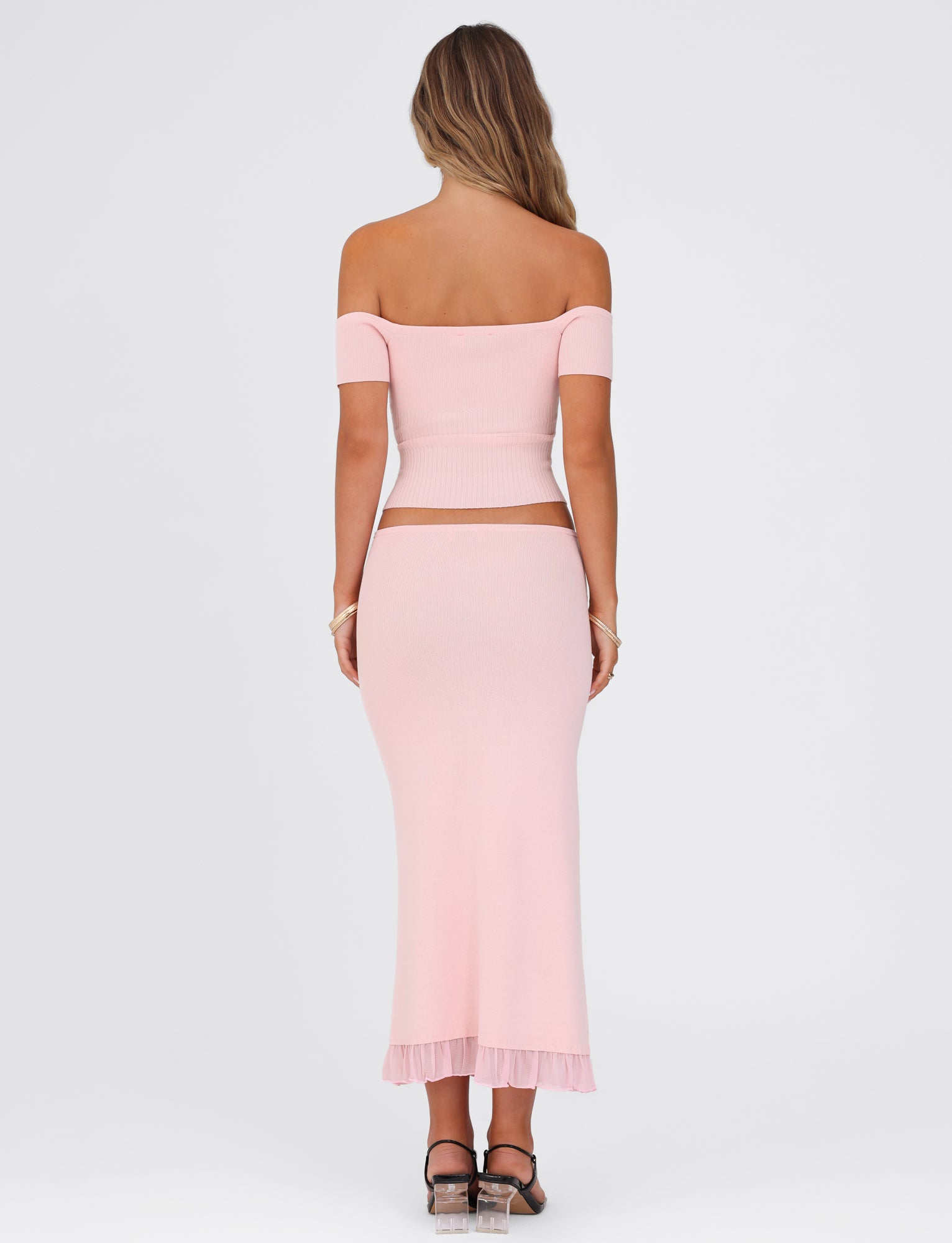 The image features the rear view of a woman wearing the LILYANA SKIRT in Baby Pink from TIGERMIST, paired with a matching off-shoulder top. She stands against a plain white background, her loose hair cascading down her back as she confidently wears black heels.