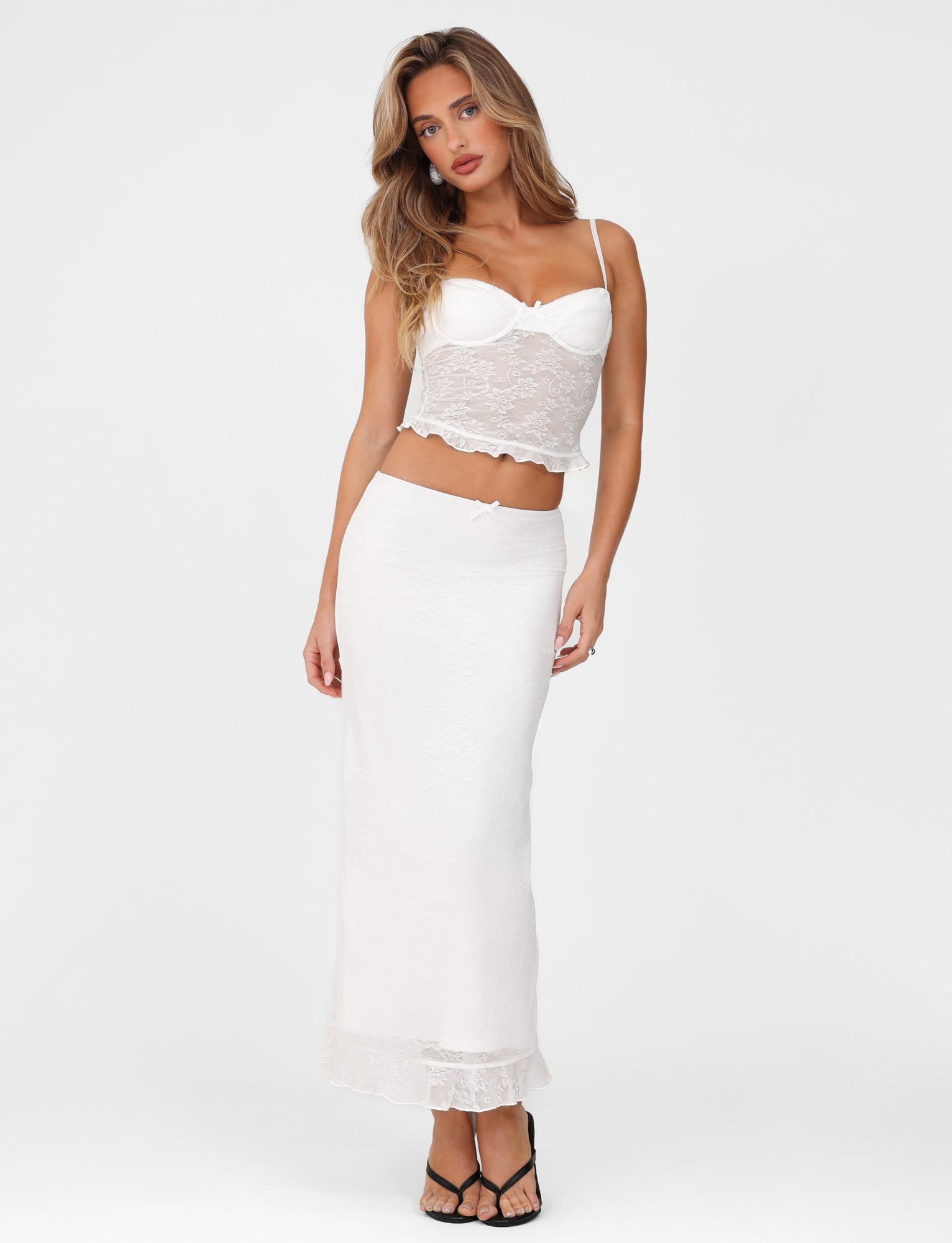 A woman wearing a white lace-trimmed camisole and the TIGERMIST Charleigh Skirt in white lace, with a low-rise waist, poses against a plain white background. She has long hair and is wearing black sandals.