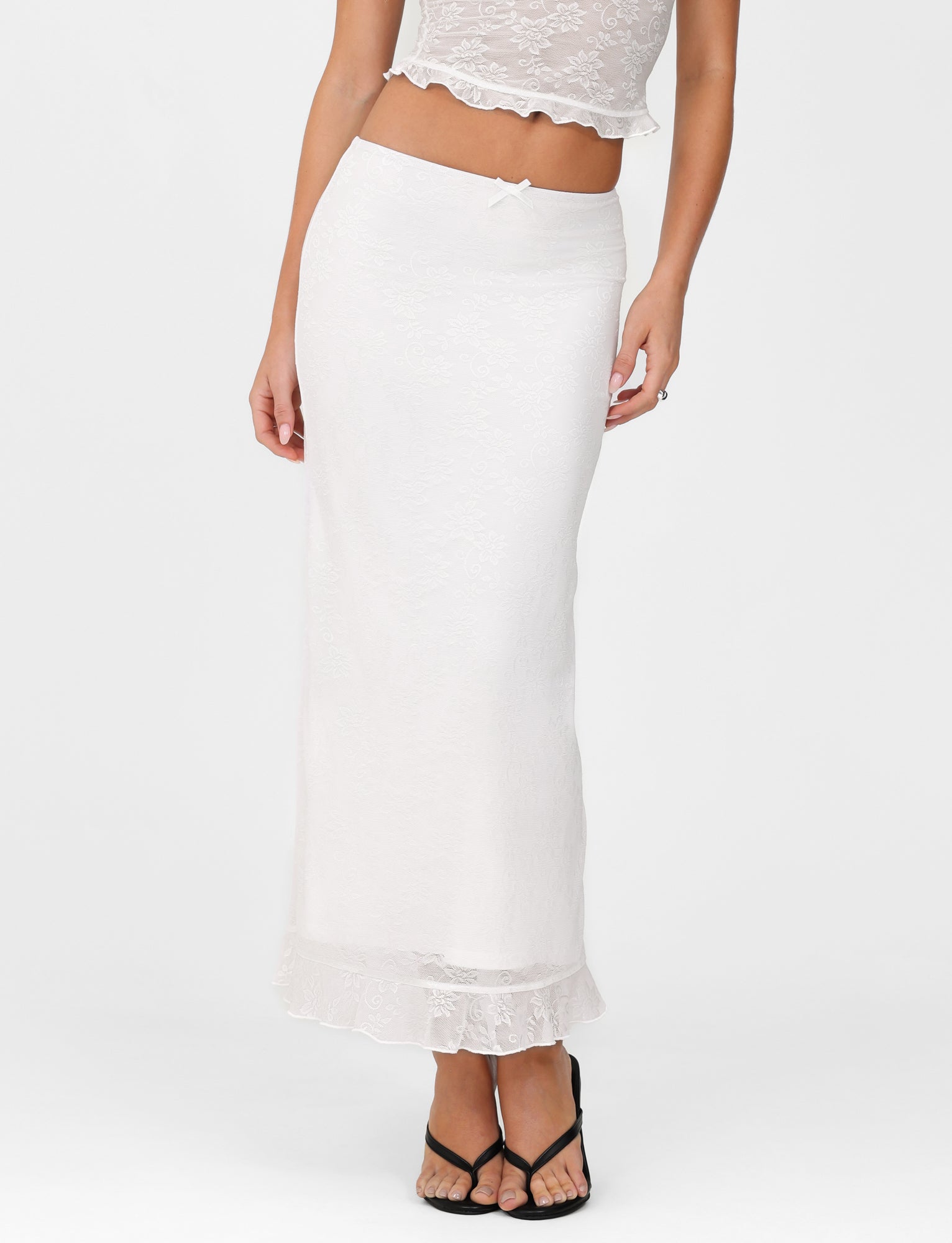 A woman wearing a white lace top and the elegant TIGERMIST CHARLEIGH SKIRT - WHITE: LACE, featuring a low-rise waist, stands against a plain background. She completes her outfit with stylish black sandals.