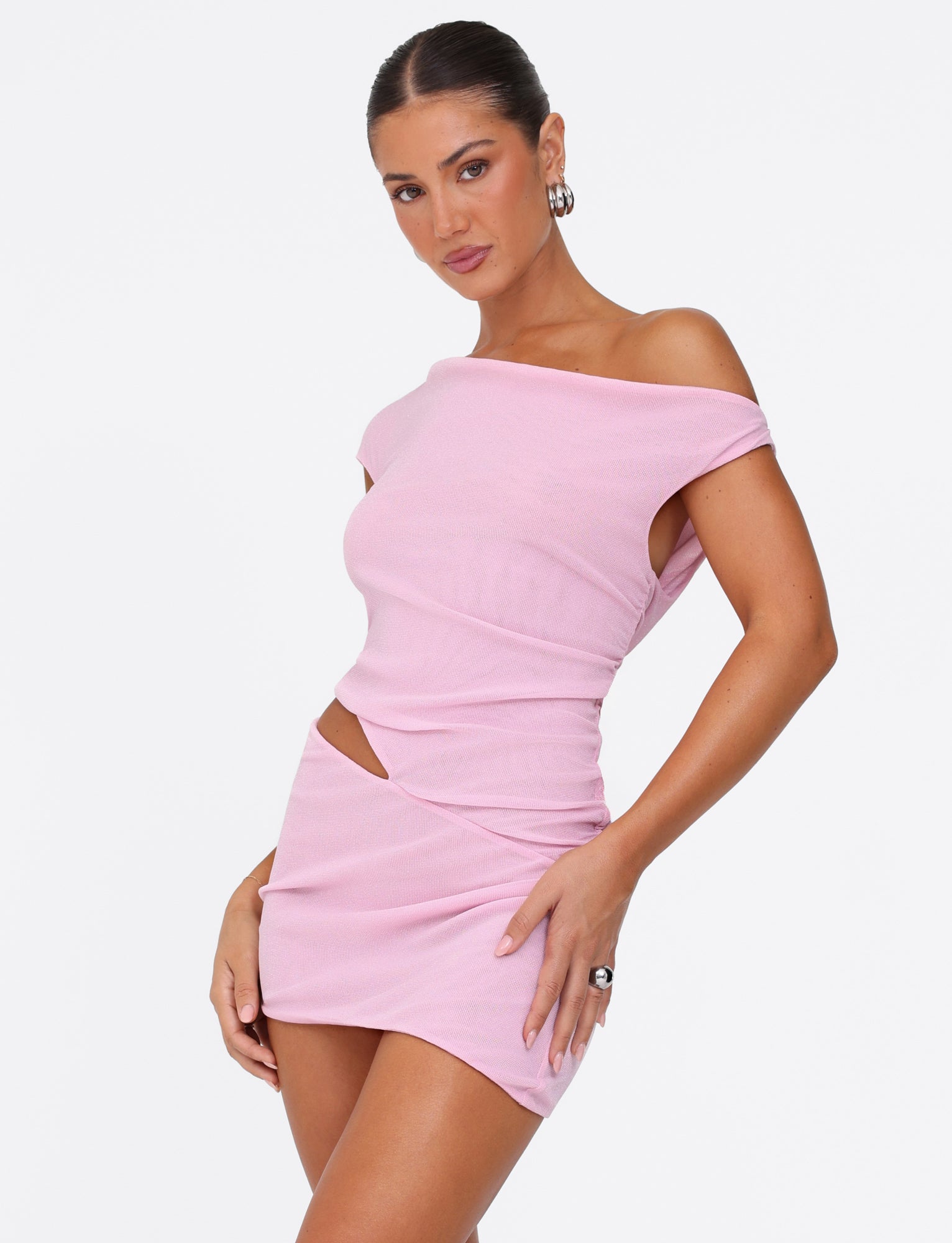 A person stands confidently in the COBI MINI DRESS 2.0 - PINK by TIGERMIST, featuring a glitter fabrication and asymmetrical design with cut-out details in baby pink. Their hair is elegantly tied back, accented by hoop earrings and a ring, all set against a plain white background. The pose exudes style and elegance.