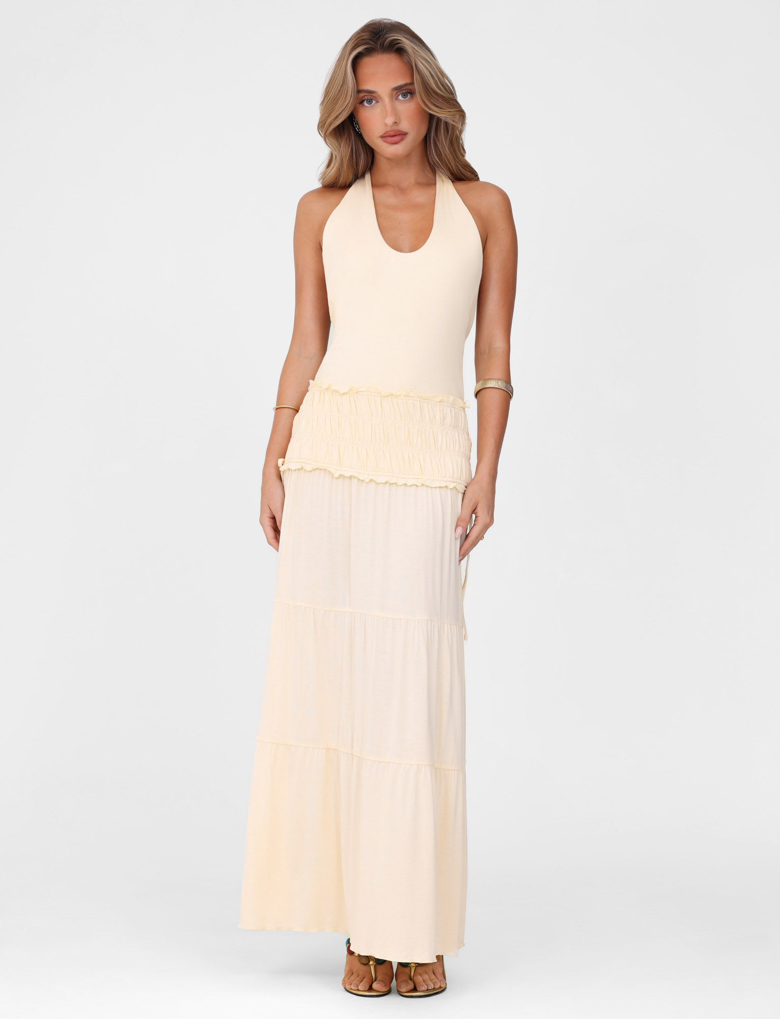 A woman with long wavy hair wears the TIGERMIST DENALI HALTER DRESS in Neutral: Bone, featuring a fitted silhouette and ruffled waist detail, standing against a plain white background and looking forward with a neutral expression.