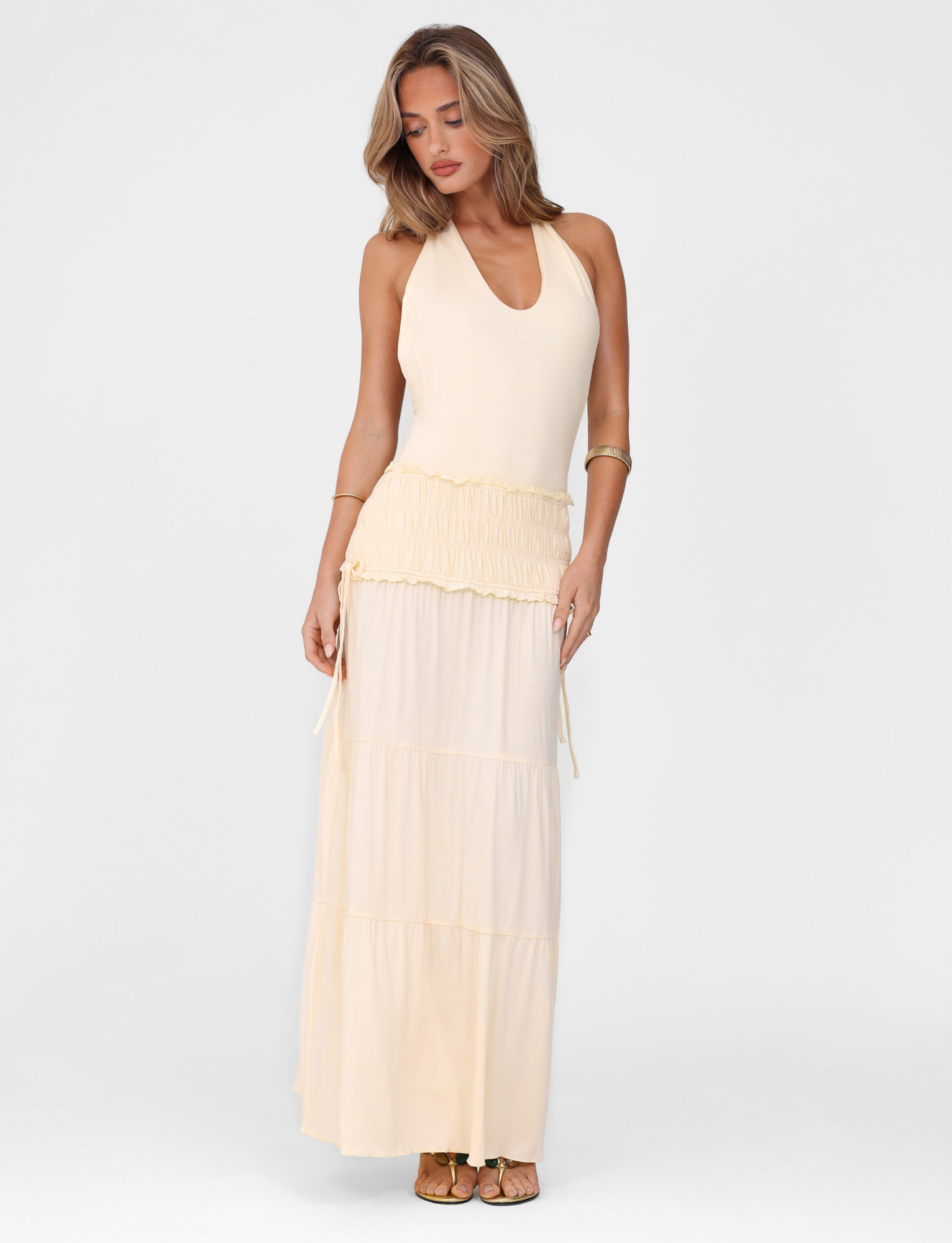 A woman models the TIGERMIST DENALI HALTER DRESS - NEUTRAL : BONE, a sleeveless maxi with a deep V-neck and tiered skirt, standing against a plain white background, looking down with one arm at her side and the other touching her waist.