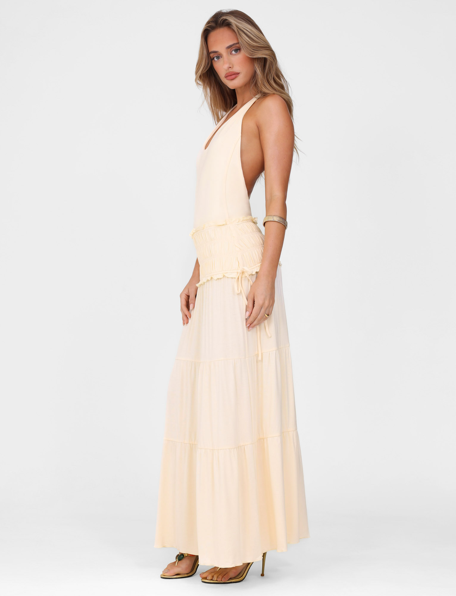 A woman with long, wavy hair wears the TIGERMIST DENALI HALTER DRESS in Neutral: Bone—a sleeveless, fitted maxi dress with a ruffled waist—paired with high heels and a gold bangle, against a plain white background.