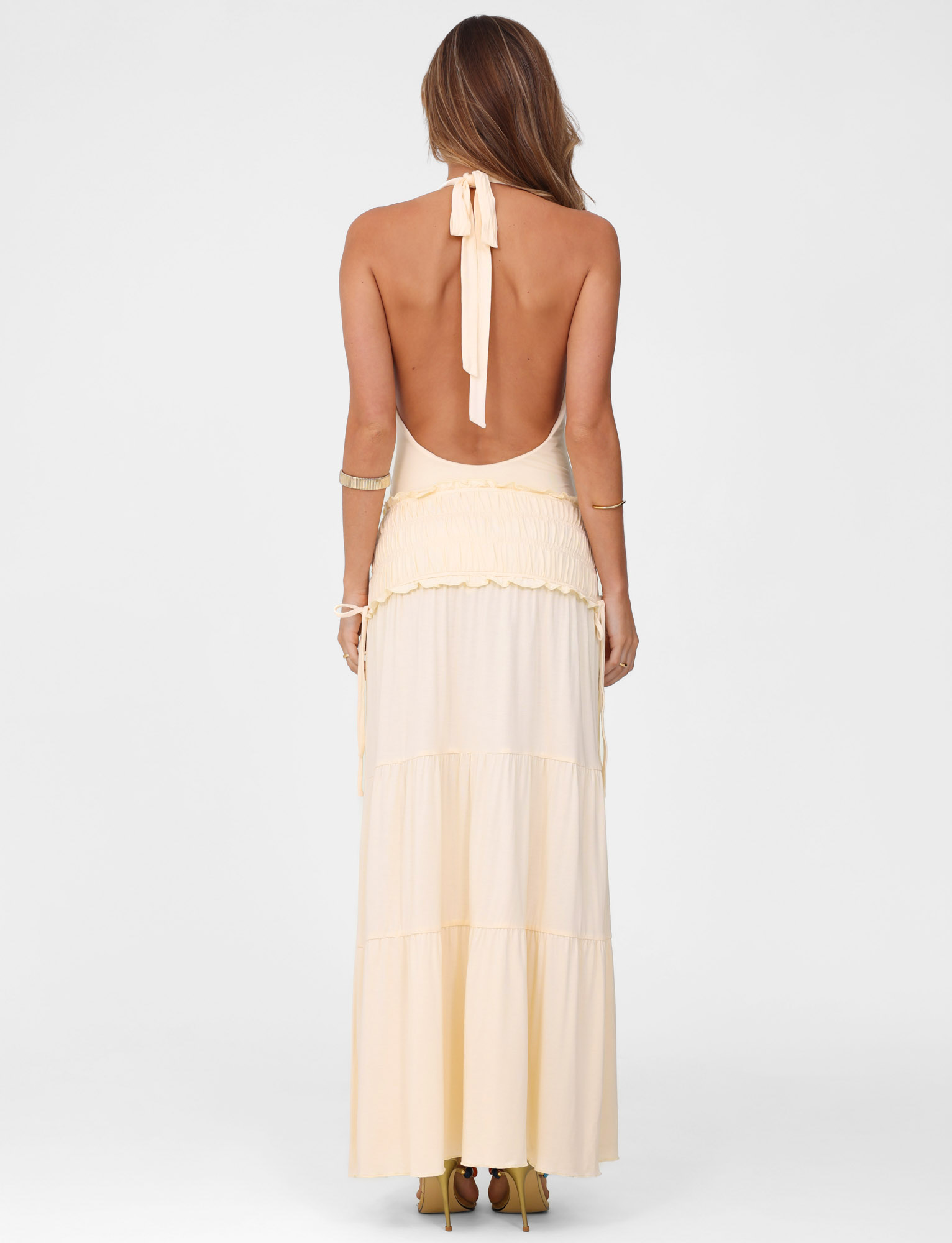 A woman faces away from the camera in the TIGERMIST DENALI HALTER DRESS - NEUTRAL : BONE, a fitted halter maxi dress. Her hair is loose, gold bangles on both arms. She stands against a plain white background.