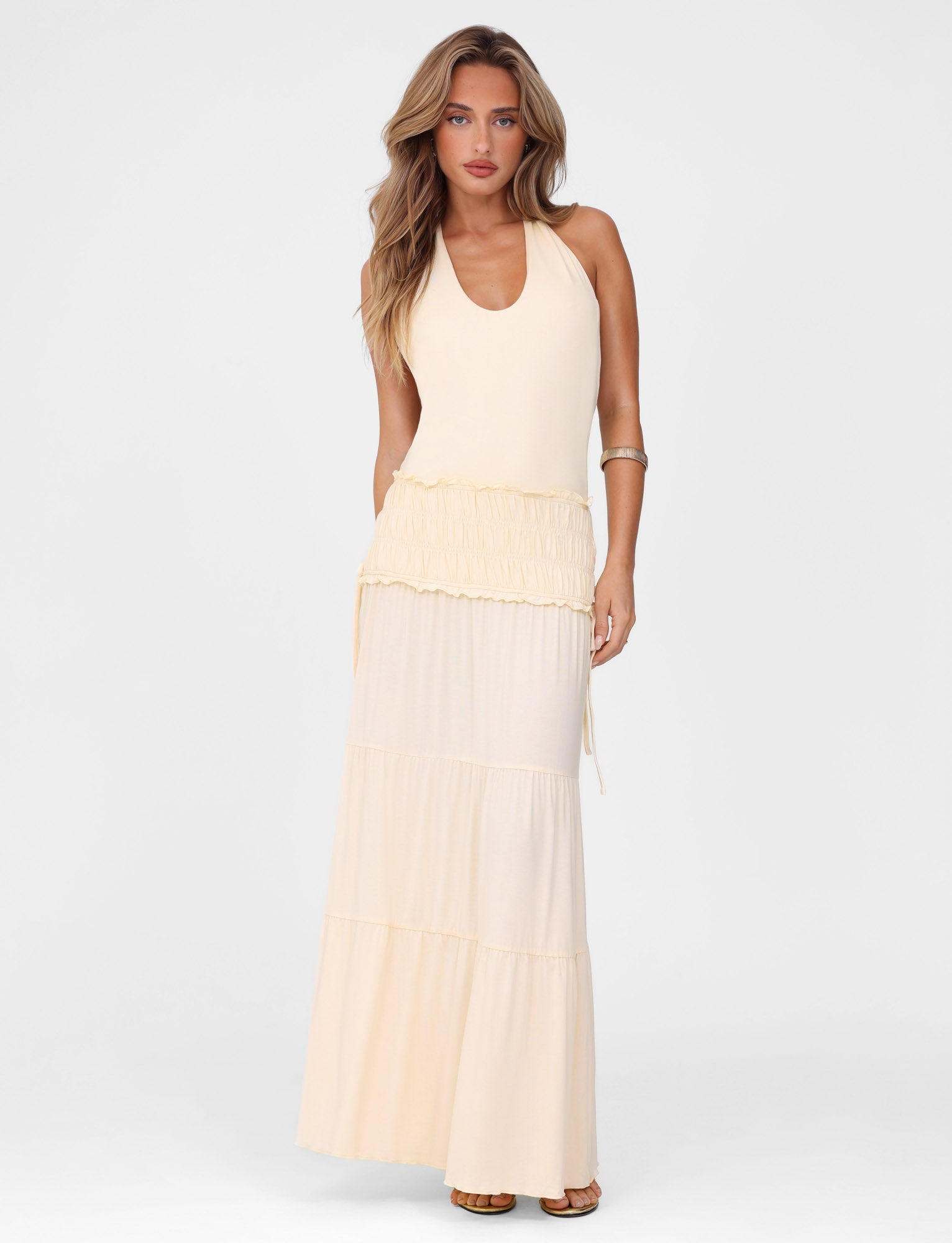 A woman with long wavy hair models the TIGERMIST DENALI HALTER DRESS in Neutral: Bone, a sleeveless maxi dress with a tiered full skirt, standing against a plain white background.