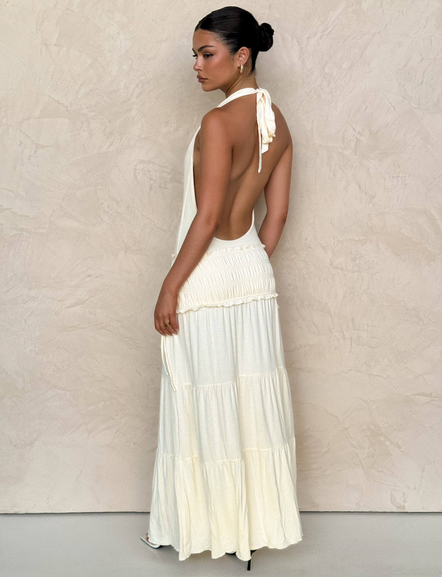 A woman stands sideways, looking back over her shoulder in the TIGERMIST DENALI HALTER DRESS - NEUTRAL: BONE, a cream-colored maxi with a fitted bodice and tiered skirt. Her hair is styled in a sleek bun against a light textured wall.