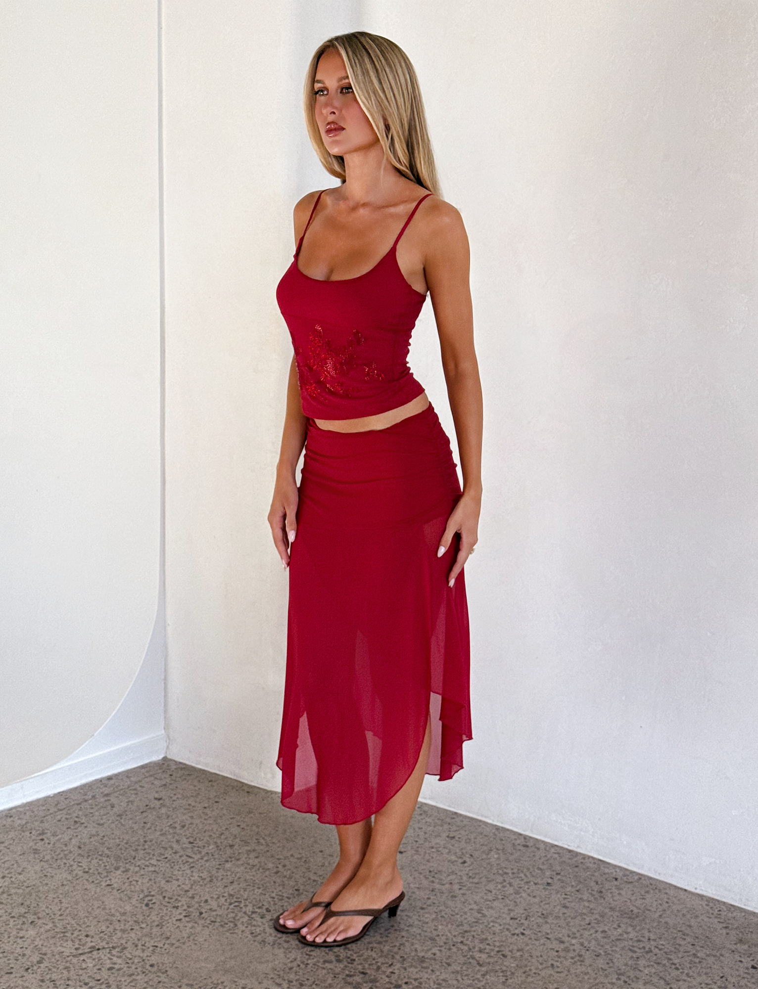 A woman stands indoors against a light wall, wearing a sleeveless red top and the TIGERMIST MARIANA SKIRT - RED with an asymmetric wrap design. Her long blonde hair flows as she completes the look with black flip-flops.