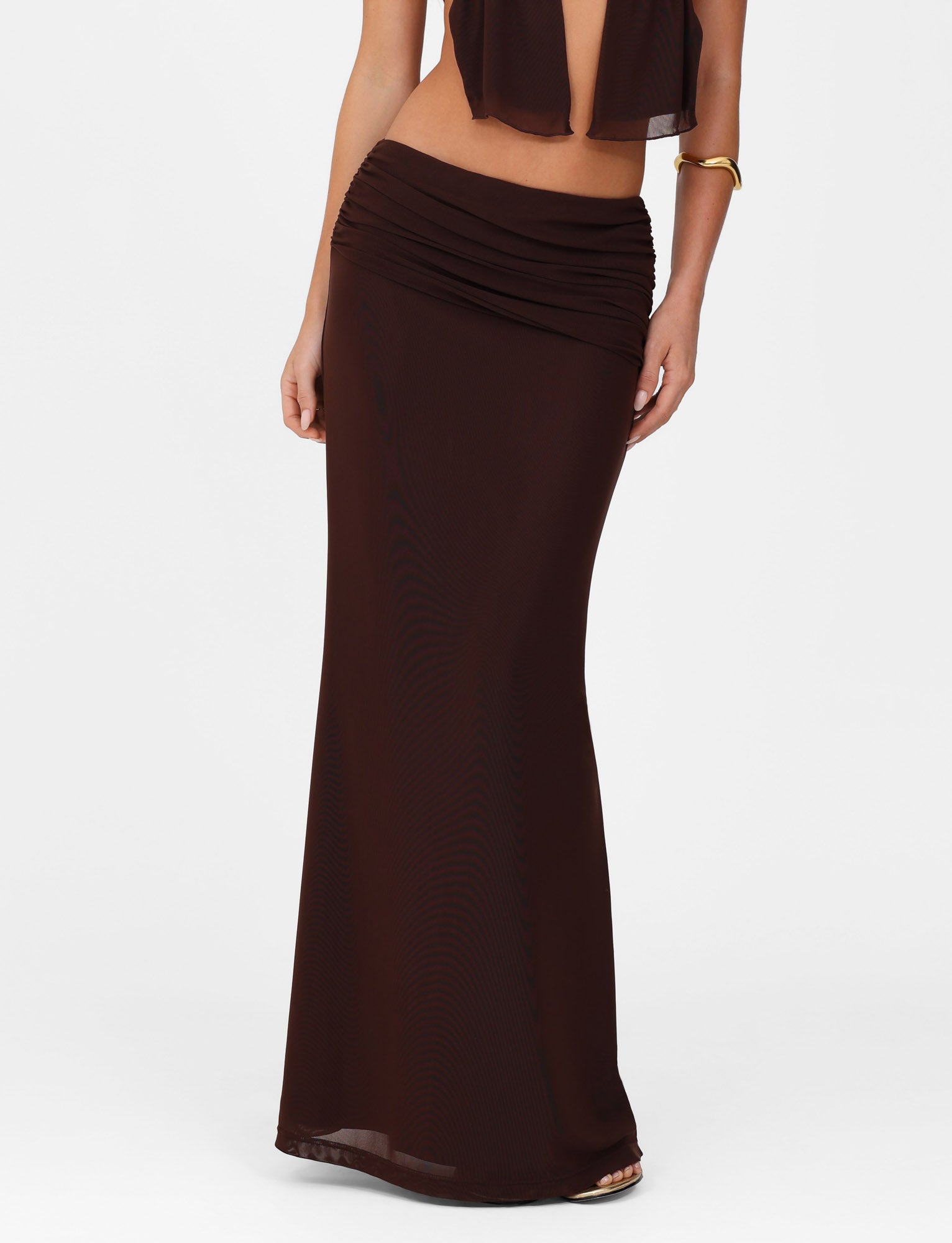A person models a chocolate brown TigerMist Esha Maxi Skirt, showcasing its gathered waist and cropped top styling. The A-line maxi skirt, fitted at the hips and slightly flared, highlights midriff exposure. A gold bangle adorns their right wrist against a white background.