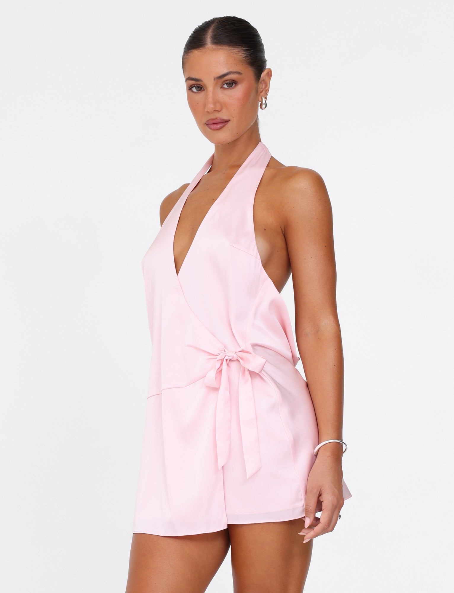 A woman is dressed in the TIGERMIST MONROE 2.0 MINI DRESS in pastel pink, echoing the elegance of a classic Monroe Dress with its halter-neck and wrap design, complete with a tied waist. Her hair is pulled back as she poses against a plain white background.