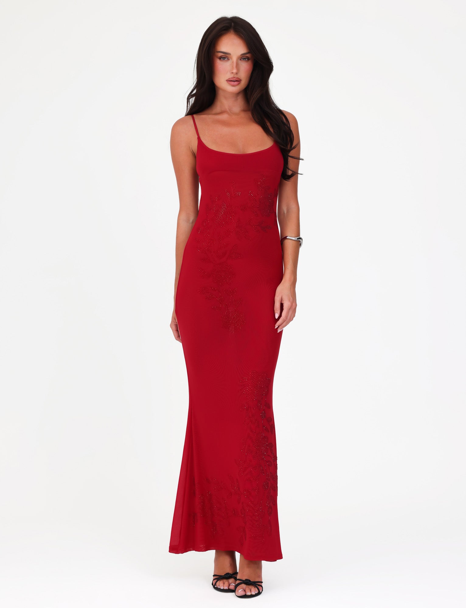 A person is dressed in a Cherry Red OCTAVIA MAXI DRESS from TIGERMIST, featuring thin straps and subtle patterns, standing against a plain white background. They have long hair and are accessorized with black sandals and a bracelet.