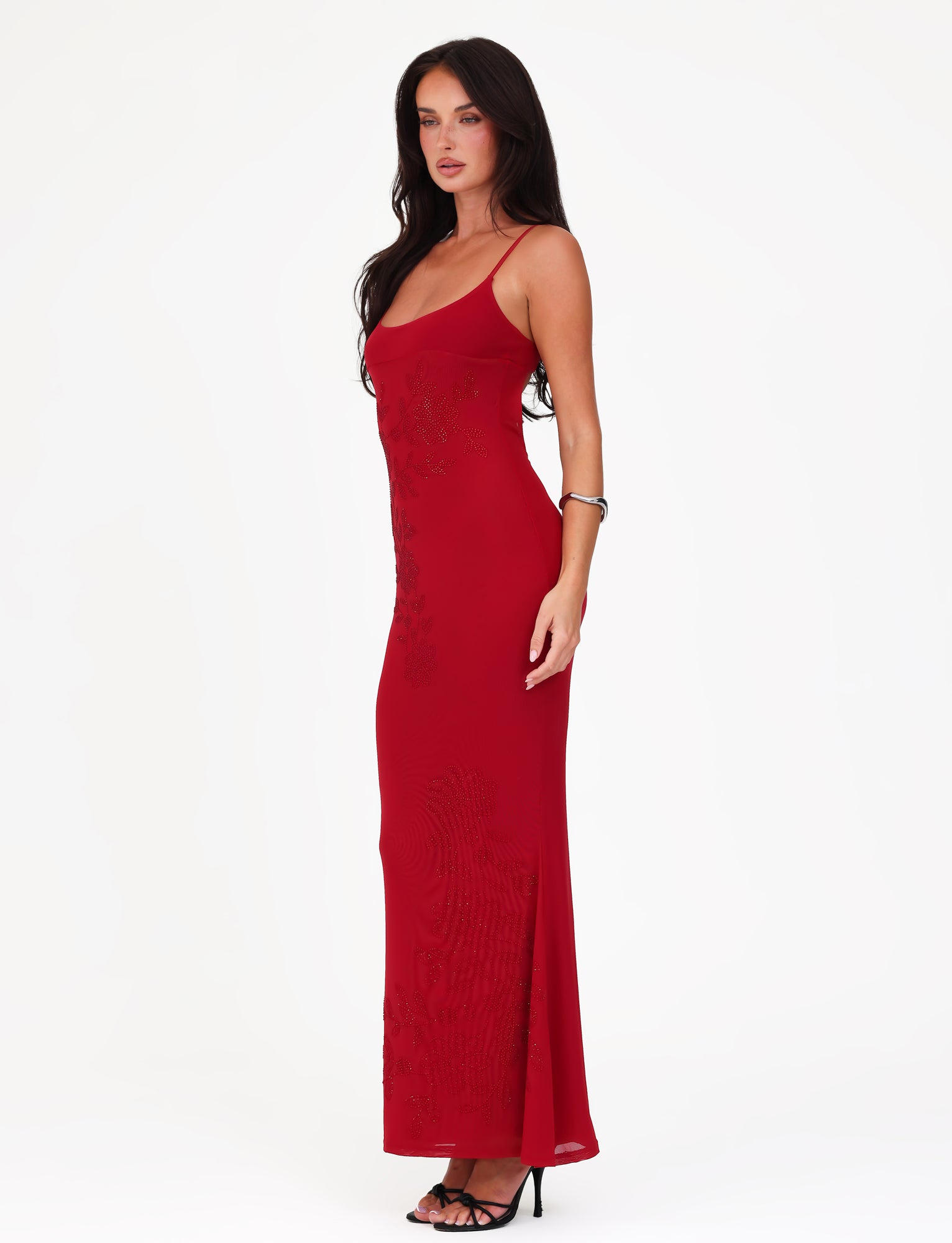 A woman in a stunning Cherry Red Octavia Maxi Dress by TIGERMIST, a long, fitted delight with thin straps, stands against a plain white background. Her long dark hair cascades down as she elegantly pairs the dress with black high heels and complements her look with a bracelet.