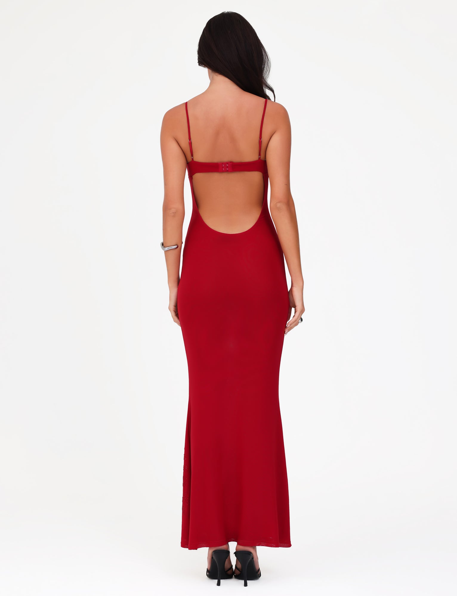 A woman, with her back to the camera, elegantly displays the exquisite OCTAVIA MAXI DRESS in Cherry Red by TIGERMIST. This backless dress boasts thin straps and delicate floral embellishments. Her long hair drapes over her shoulders, while black high-heeled shoes complete her ensemble against a plain white backdrop.