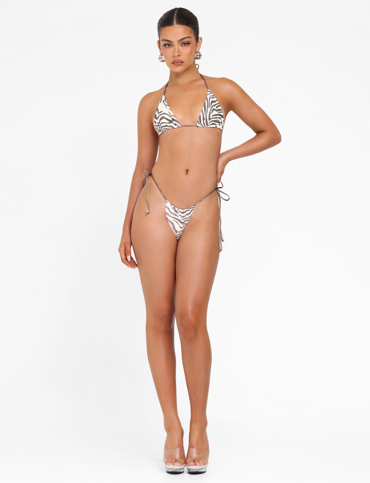 A woman stands against a white background wearing the TIGERMIST AVE BIKINI TOP - NEUTRAL : ZEBRA with true-tie halter neck and unpadded triangle cups, styled with clear high heels. She rests one hand on her hip and keeps a neutral expression.