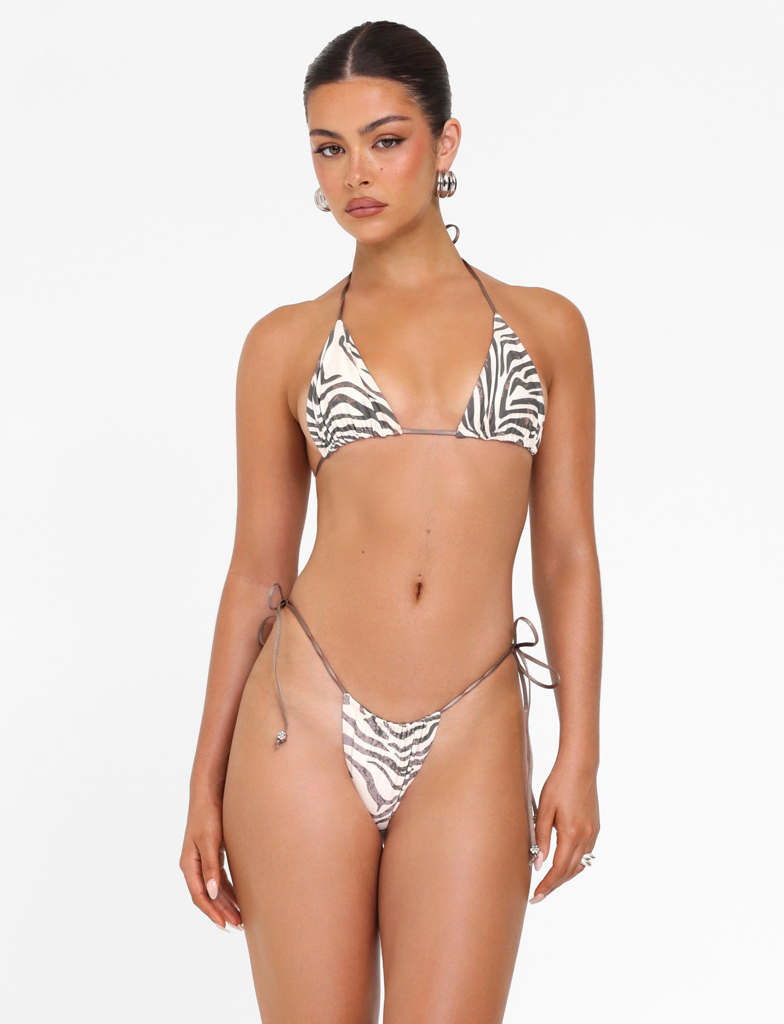 A woman wears the TIGERMIST AVE BIKINI TOP - NEUTRAL : ZEBRA, featuring a true-tie halter neck and unpadded triangle cups, standing against a white background with her hair styled back and minimal accessories.