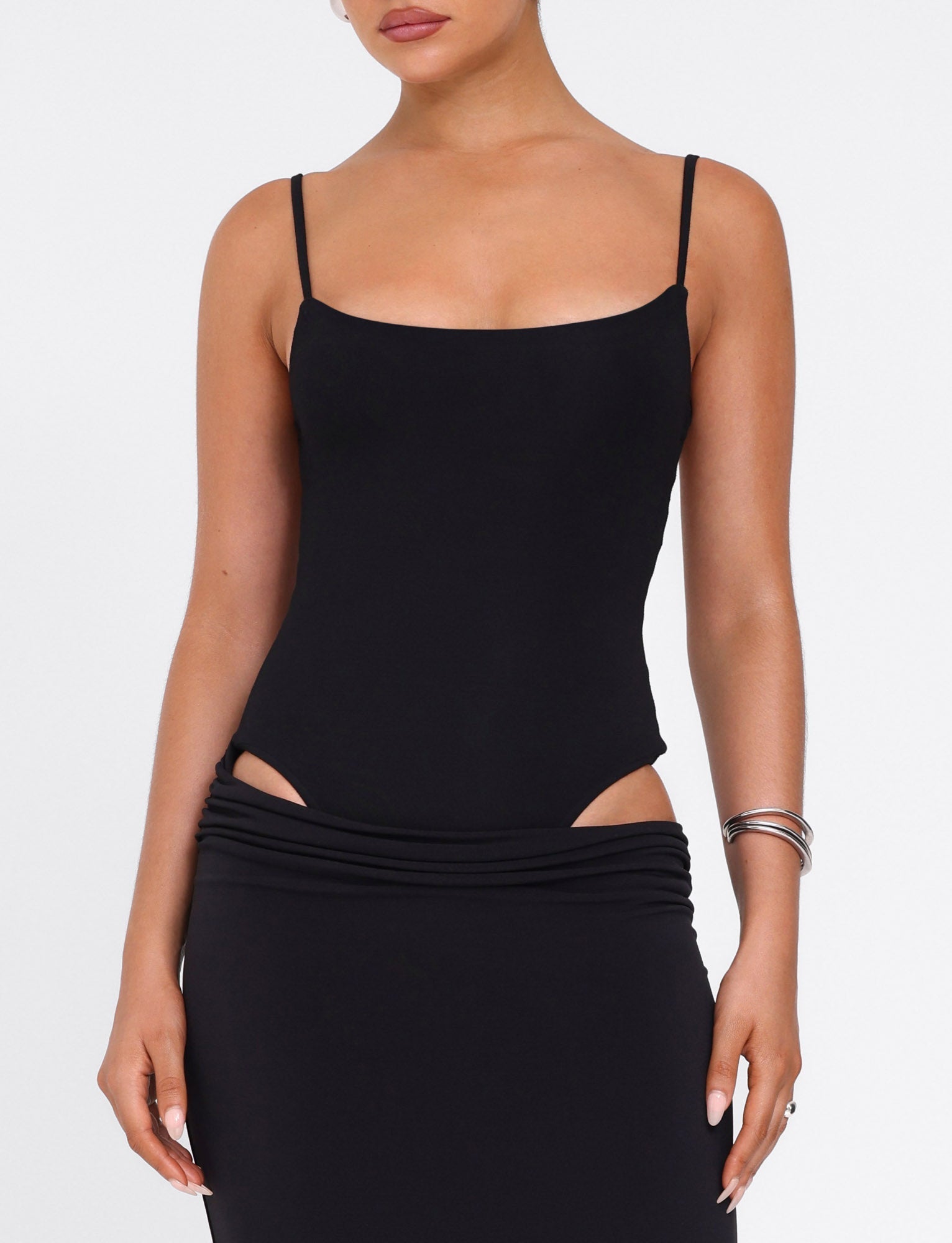 A woman models the TIGERMIST UMA DRESS - BLACK, a sleeveless bodycon dress with adjustable straps and side cutouts, accessorized with silver bangles, against a plain white background.