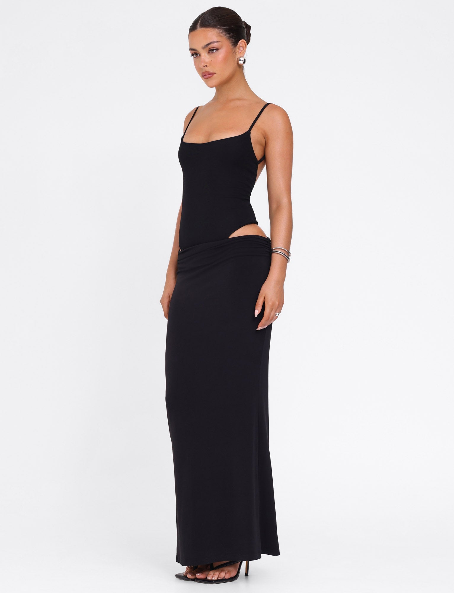 A woman models the TIGERMIST UMA DRESS - BLACK, a sleeveless, form-fitting maxi dress with adjustable straps and hip cut-out details. She pairs it with high heels and silver jewelry, hair sleekly pulled back, set against a white background.