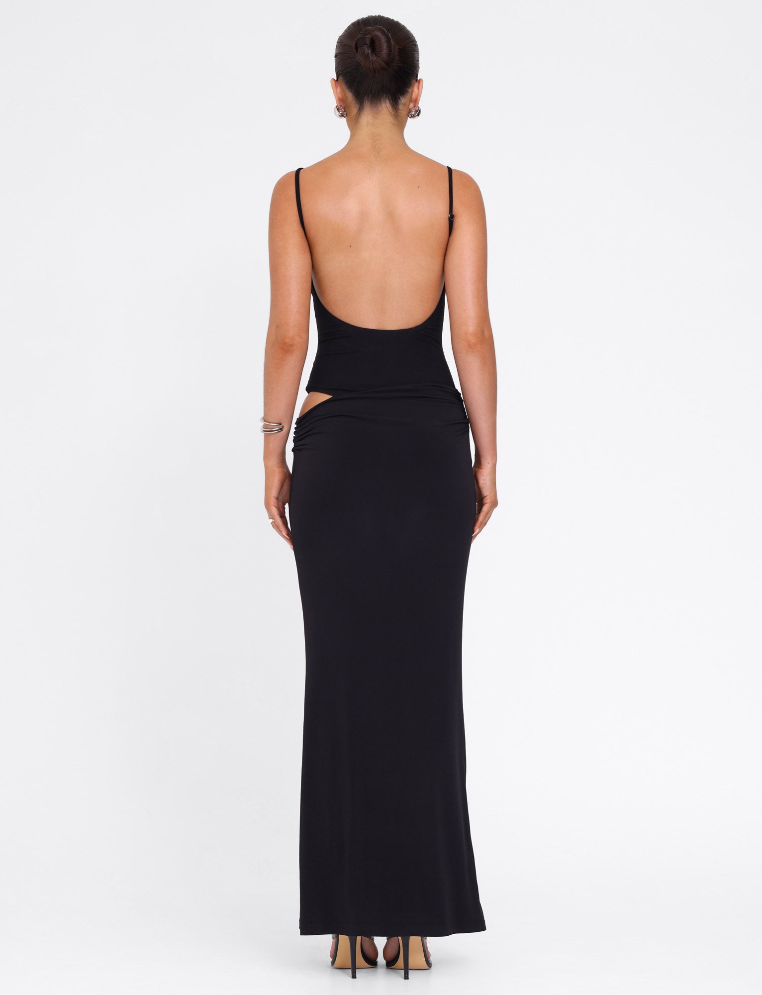 A model faces away, wearing the TIGERMIST UMA DRESS in black, featuring adjustable straps and waist cut-outs. Hair is in a neat bun, paired with high heels against a plain white backdrop.