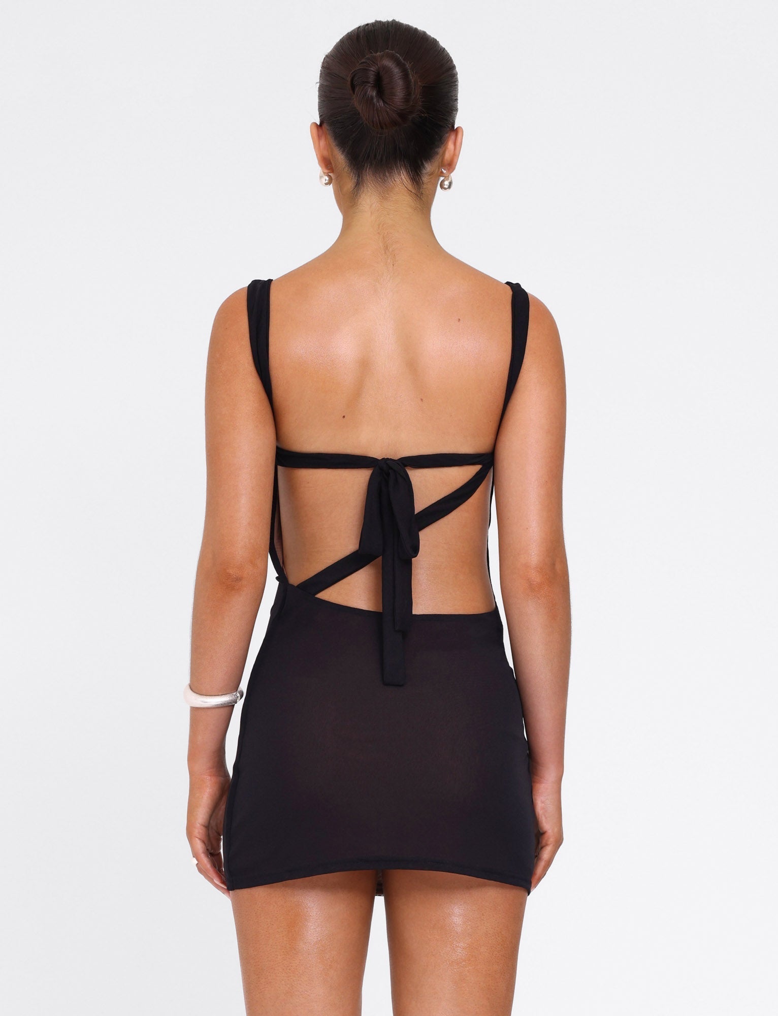 A woman faces away wearing the TIGERMIST FREYA DRESS in black, a buttery soft mini dress with an open back and tie details. Her hair is in a neat bun, accessorized with small hoops and a white bangle on her left wrist.