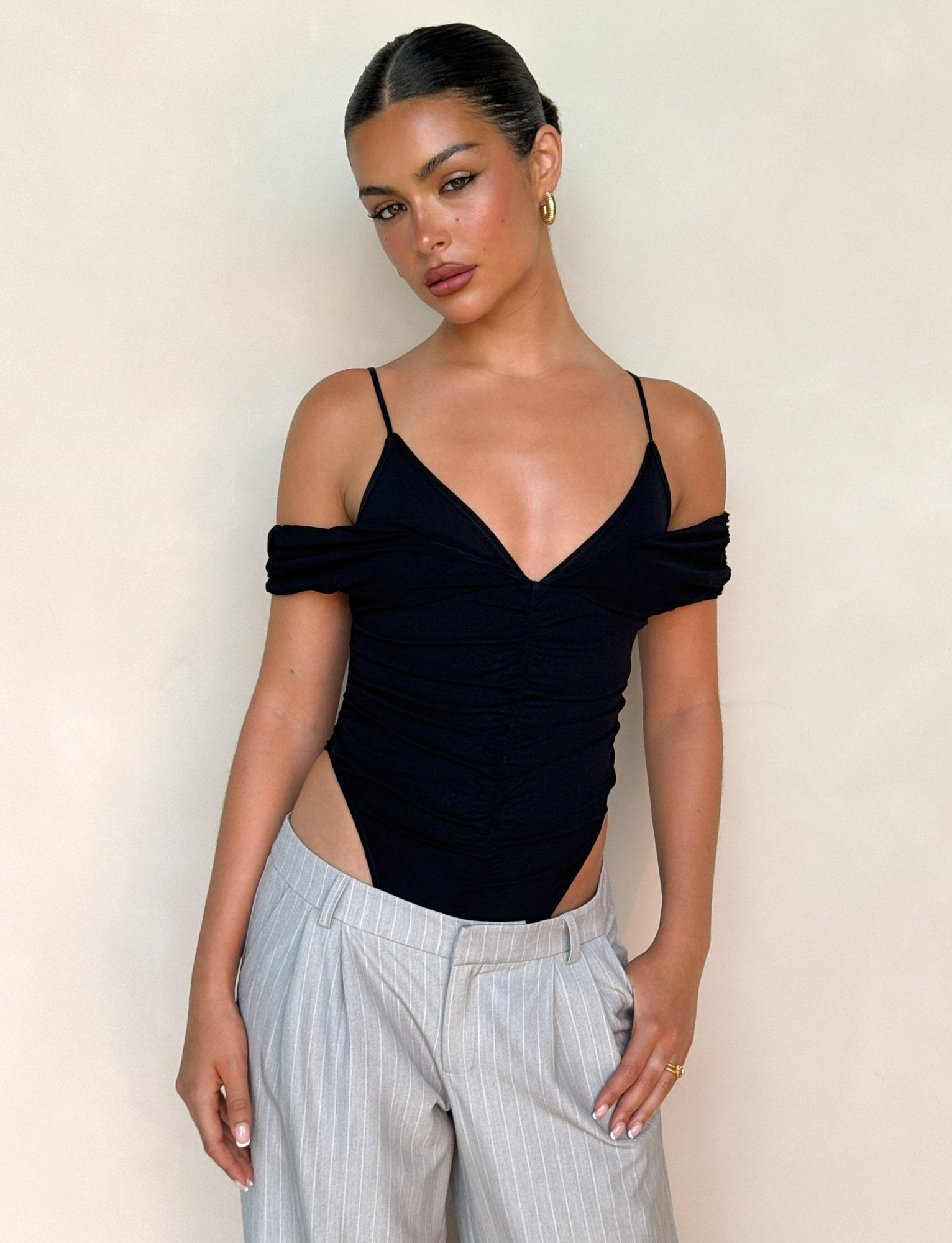 A woman with slicked-back hair models the TIGERMIST DRAYA BODYSUIT in black and light gray pinstripe trousers, standing against a plain beige background and gazing at the camera with a relaxed expression.