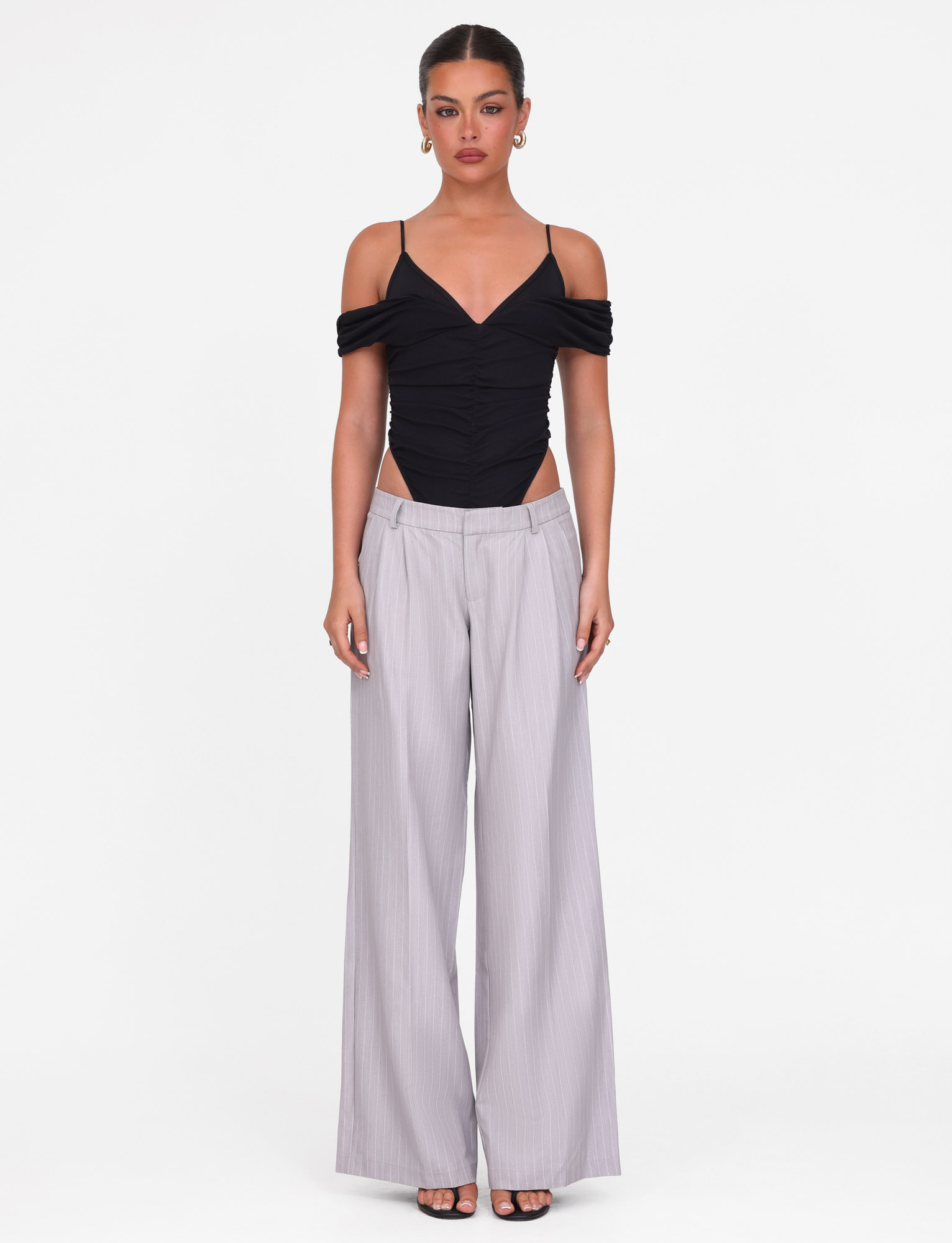A woman faces forward wearing the TIGERMIST DRAYA BODYSUIT in black, featuring an off-shoulder mesh design with thin straps, paired with light gray wide-leg trousers. Her hair is pulled back and she wears earrings against a plain white background.