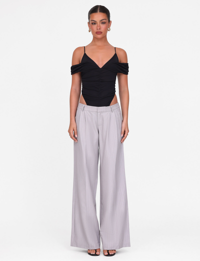 A woman faces forward wearing the TIGERMIST DRAYA BODYSUIT in black, featuring an off-shoulder mesh design with thin straps, paired with light gray wide-leg trousers. Her hair is pulled back and she wears earrings against a plain white background.