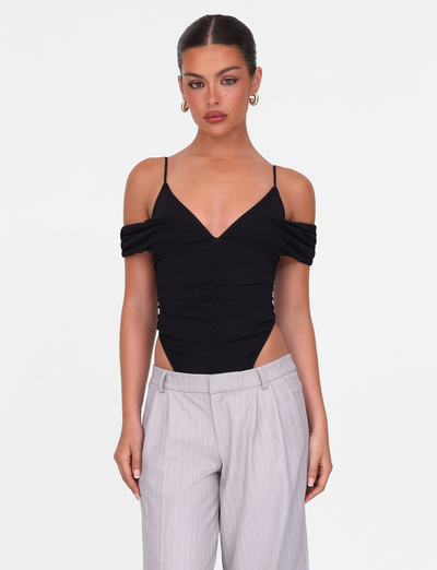 A woman with dark hair in a sleek bun wears the DRAYA BODYSUIT - BLACK by TIGERMIST, an off-shoulder style with thin straps, paired with light gray pinstripe trousers as she stands facing forward against a white background.