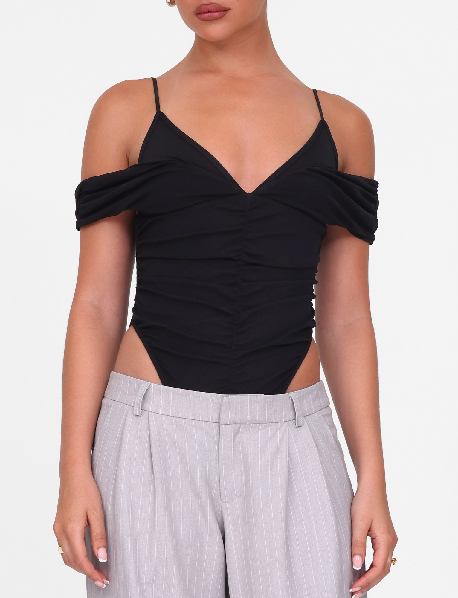 A woman models the TIGERMIST DRAYA BODYSUIT - BLACK, an off-shoulder, bodycon mesh piece with thin straps, styled with light gray pinstriped pants. The photo is cropped from shoulders to hips against a white background.