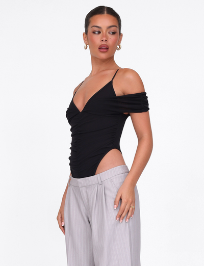 A woman wears the TIGERMIST DRAYA BODYSUIT in black, a chic off-shoulder bodycon piece with thin straps, paired with high-waisted light gray pinstripe pants. She has her hair tied back, hoop earrings on, gazing to the side.