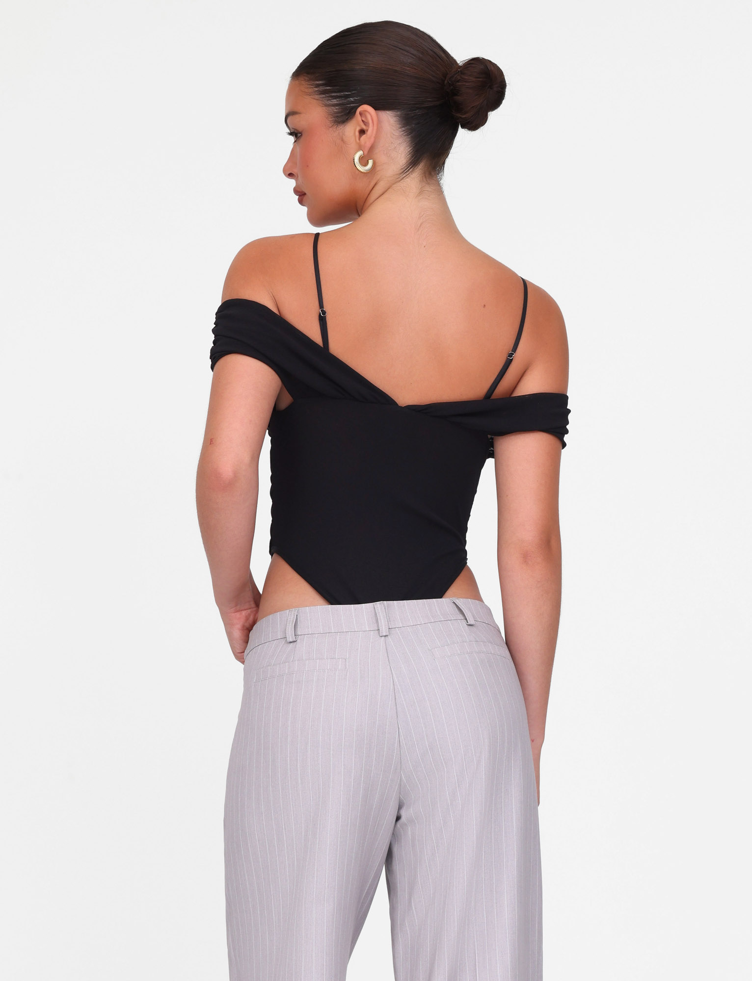 A woman with her hair in a bun, facing away from the camera, wears the TIGERMIST DRAYA BODYSUIT in black with spaghetti straps and light gray high-waisted trousers. The background is plain white.