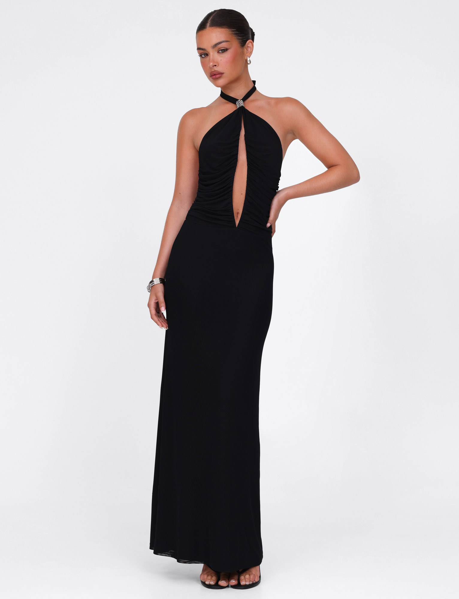 A woman stands against a white background wearing the TIGERMIST ZARA MAXI DRESS in black—a sleeveless halter style with front keyhole cutout and high-stretch, figure-flattering fabric. She accessorizes with earrings, a bracelet, and hair styled back.