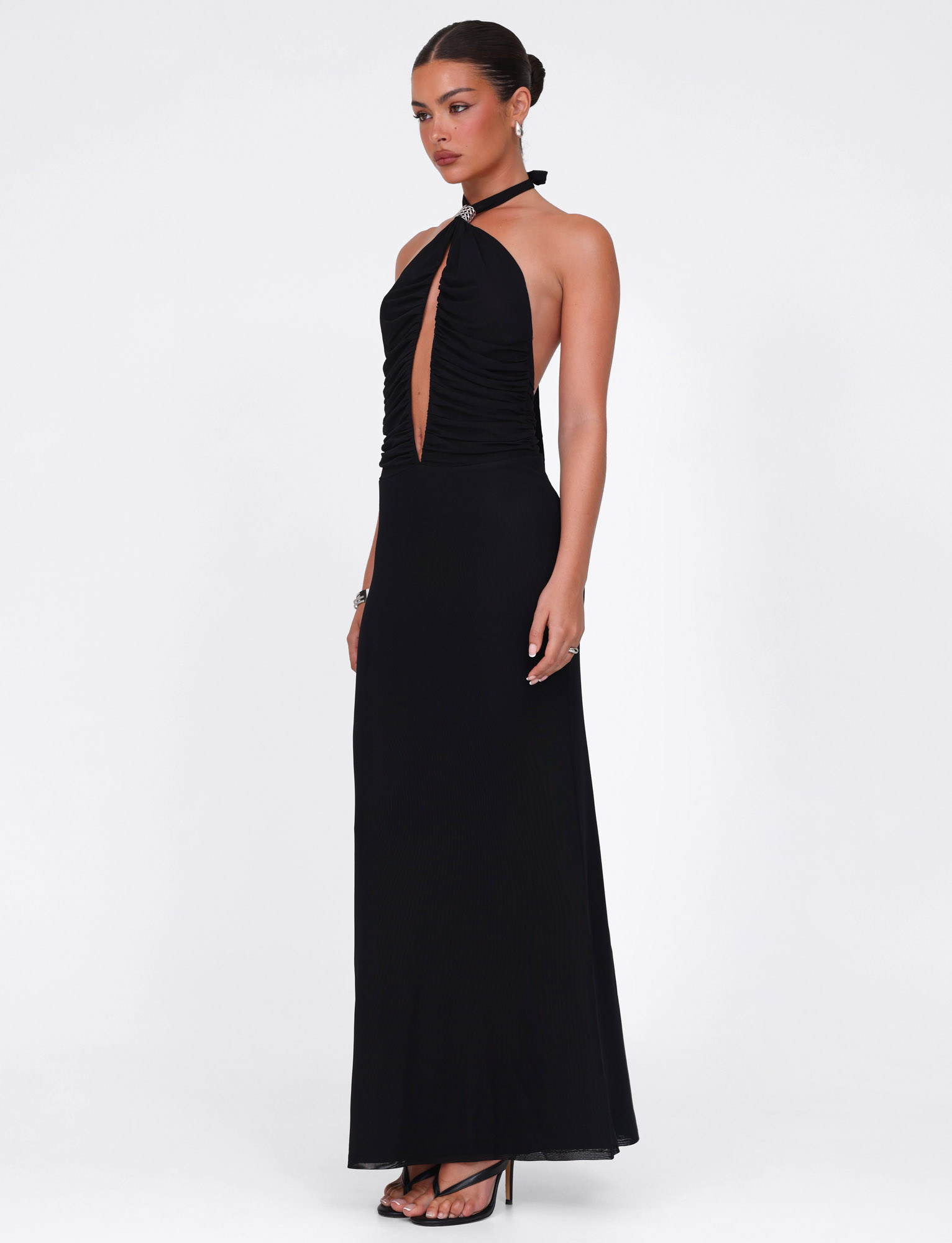 A woman stands against a white background wearing the TIGERMIST ZARA MAXI DRESS in black, a sleeveless halter style with a front keyhole cutout and high-stretch fabric, paired with black high-heeled sandals and a sleek bun.