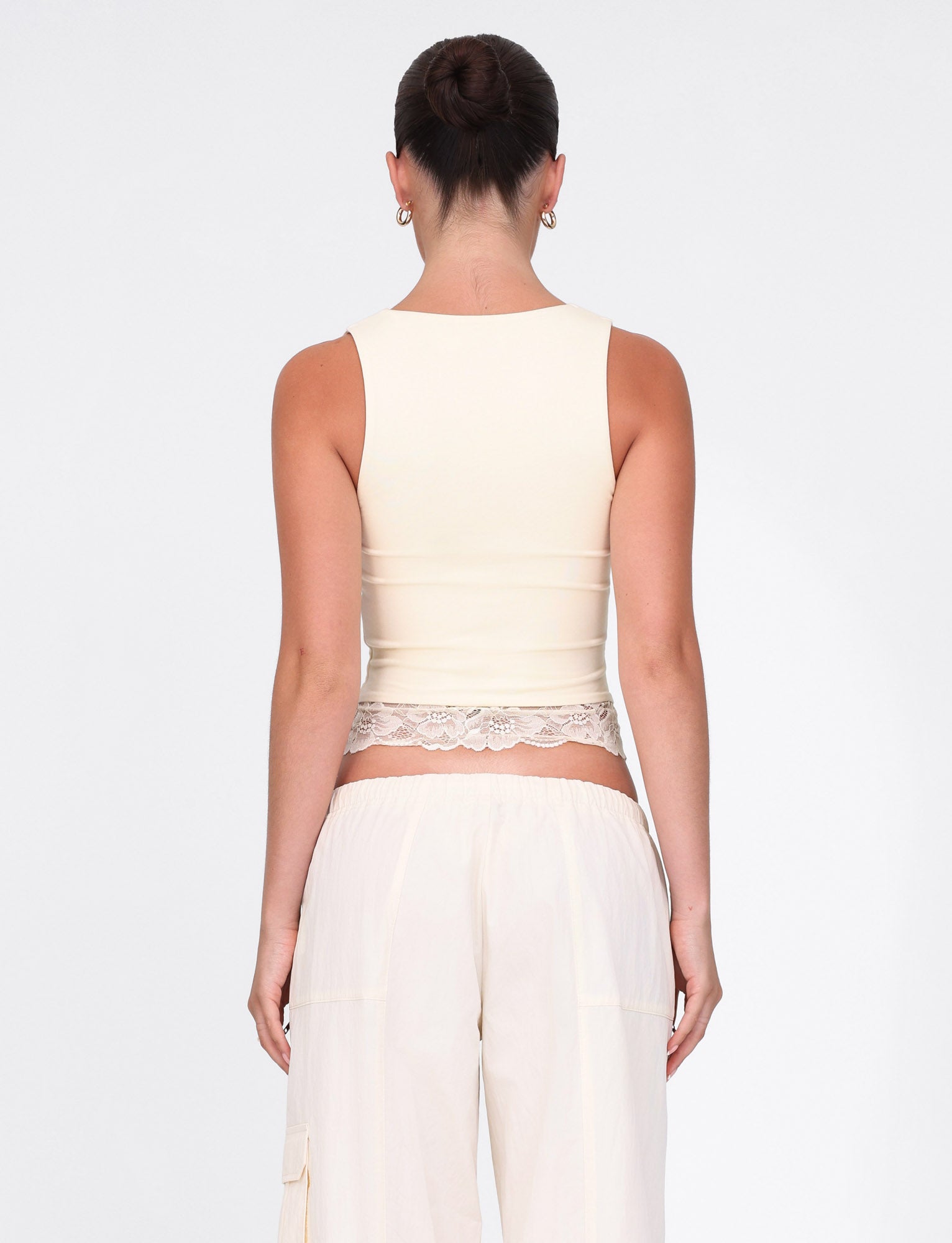 A woman with her hair in a bun showcases the TIGERMIST DIONE LACE TOP in Soft Butter, complemented by off-white pants, creating an elegant look against a plain white background.