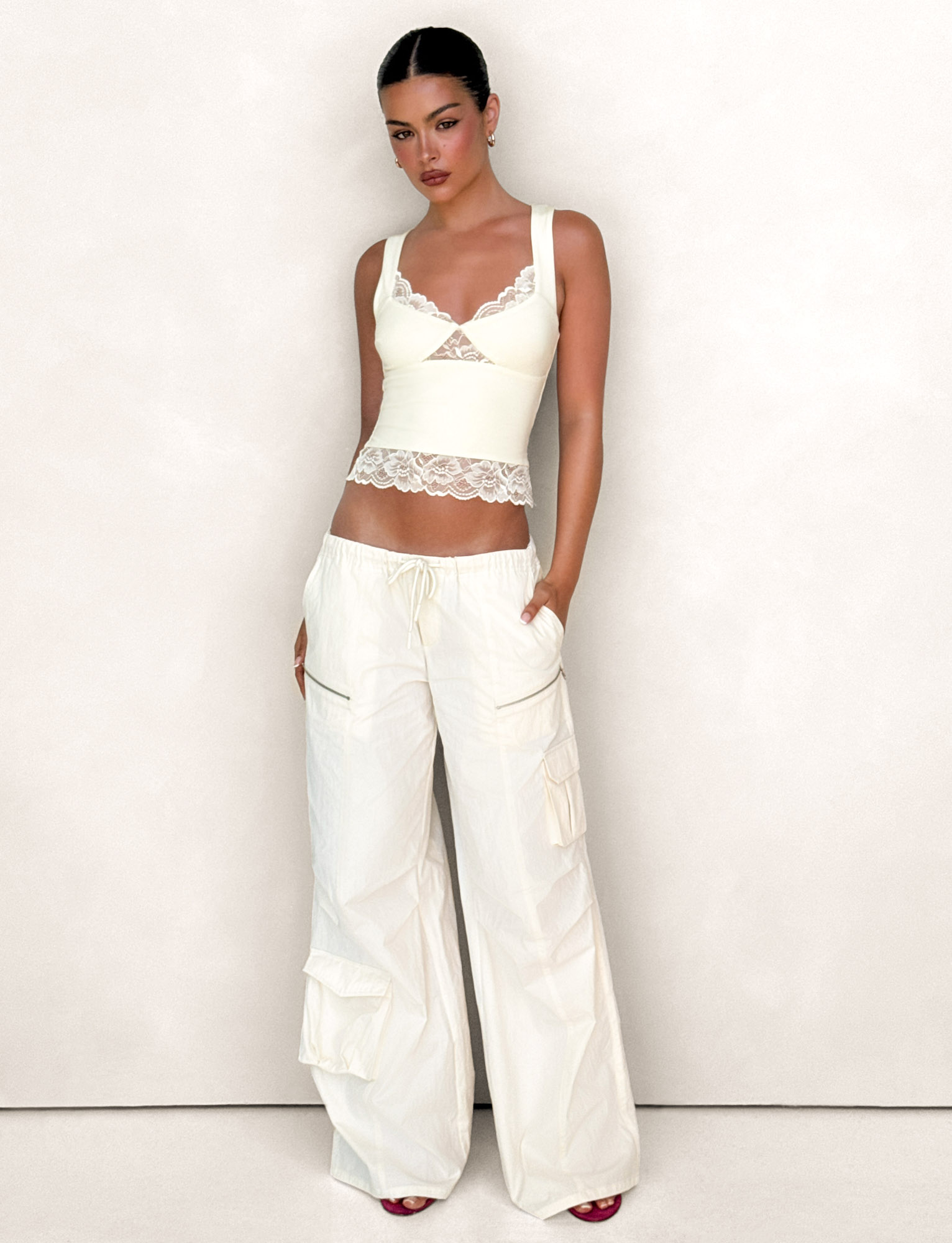 A woman poses confidently with slicked-back dark hair, wearing the TIGERMIST HERMIONE PANT in Soft Butter Yellow—stylish wide-leg cargo pants with a drawstring waist and side pockets—paired with a fitted ivory lace-trim tank top.
