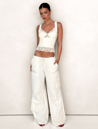 A woman poses confidently with slicked-back dark hair, wearing the TIGERMIST HERMIONE PANT in Soft Butter Yellow—stylish wide-leg cargo pants with a drawstring waist and side pockets—paired with a fitted ivory lace-trim tank top.