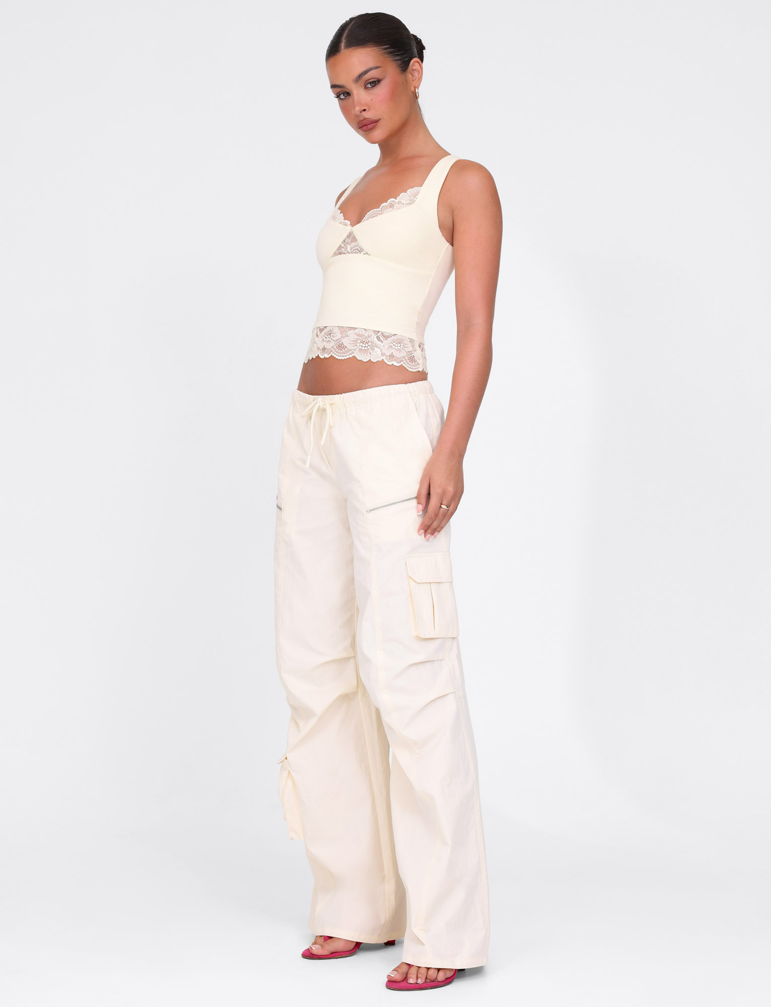 A woman poses confidently against a white background wearing TIGERMIST’s HERMIONE PANT in Soft Butter, paired with a cream lace-trimmed crop top and pink heeled sandals.