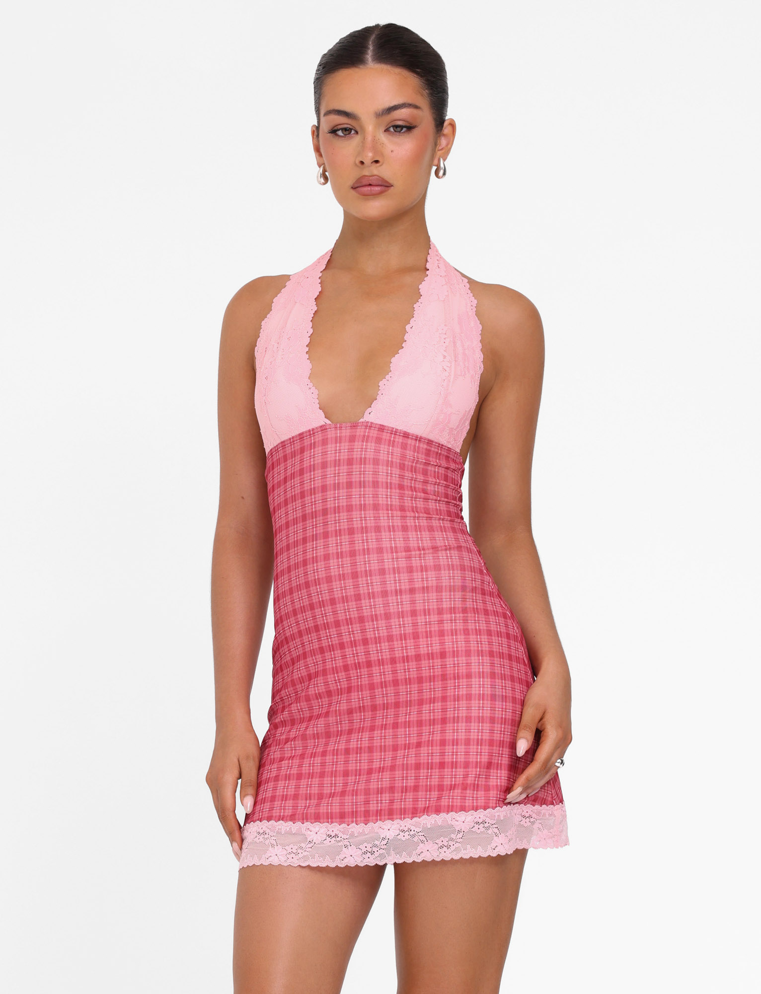 A woman stands against a white background wearing the TIGERMIST NAVIA DRESS - RED: RED CHECK, a pink halter-neck mini dress with lace-trimmed deep neckline and hem, featuring a red plaid skirt. Her hair is tied back as she looks forward.