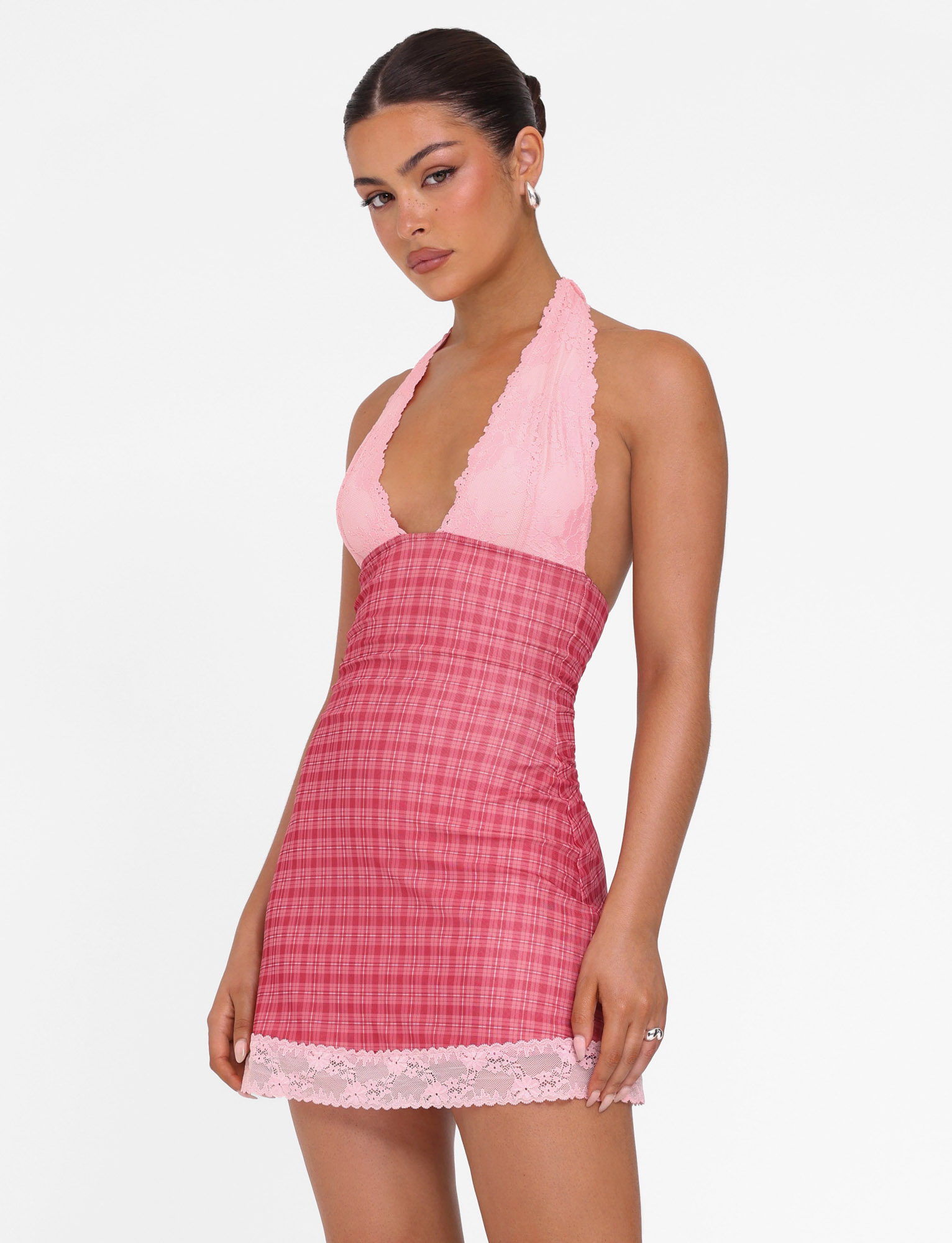 A woman poses against a white background, wearing the TIGERMIST NAVIA DRESS - RED CHECK: a sleeveless pink halter dress with a lace-trimmed neckline and red plaid skirt finished with a lace hem.