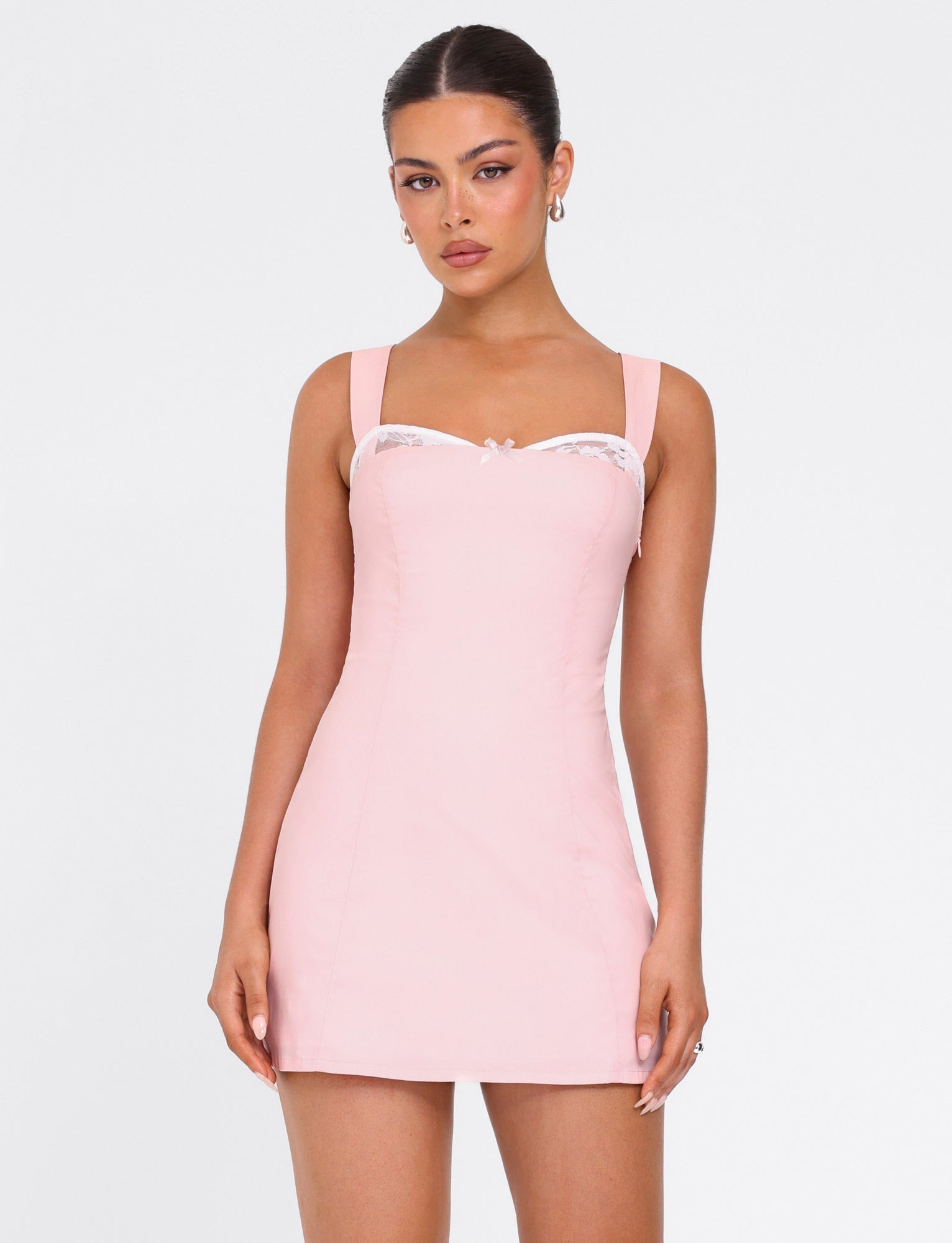 A woman models the TIGERMIST COLSON MINI DRESS in baby pink, featuring wide straps and a lace-trimmed neckline, as she stands against a plain white background and looks at the camera.