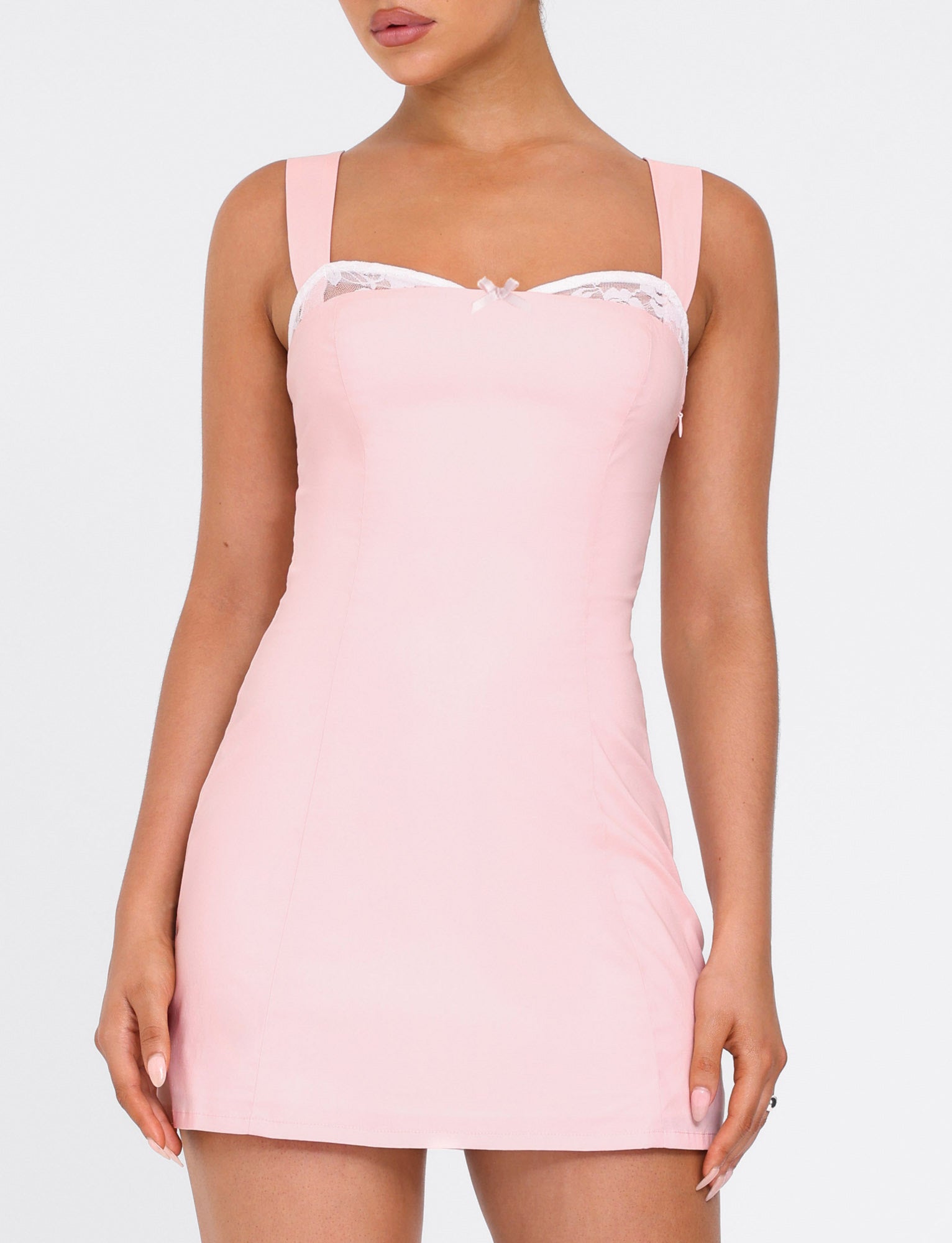 A woman wears the TIGERMIST COLSON MINI DRESS - BABY PINK, a form-fitting light pink mini dress with wide shoulder straps and white lace trim along the top, against a plain white background.